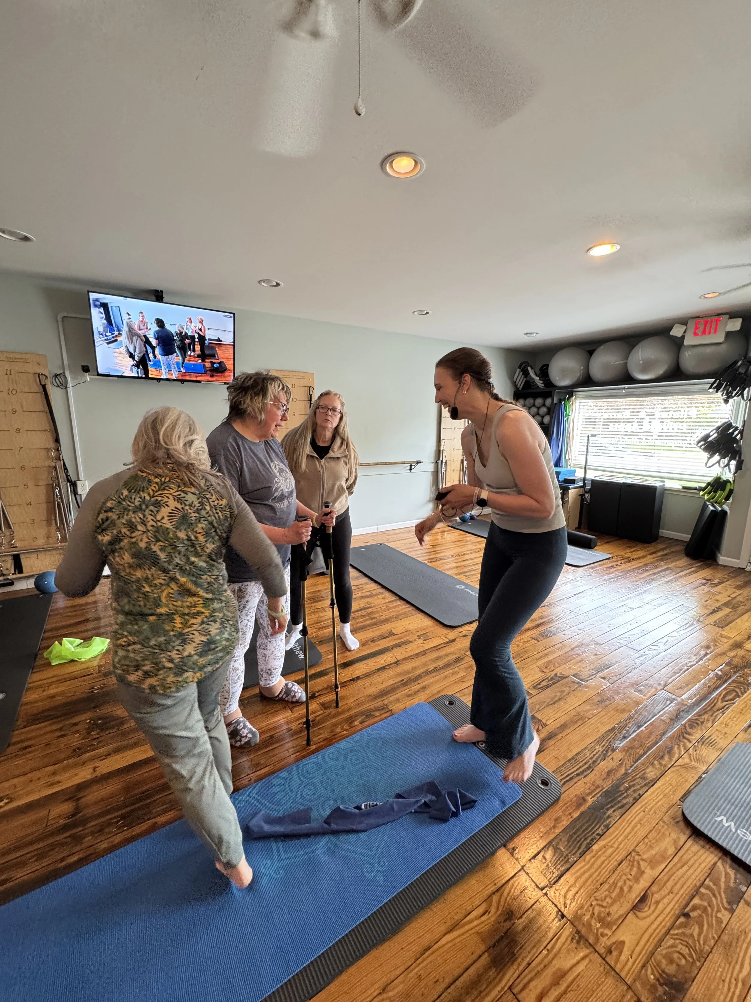 A fitness instructor leading a yoga class in a gym. Three women are participating in the yoga session, standing on mats and holding yoga props. The instructor, wearing a headset microphone, is demonstrating a pose. The gym has a wooden floor, wall-mo