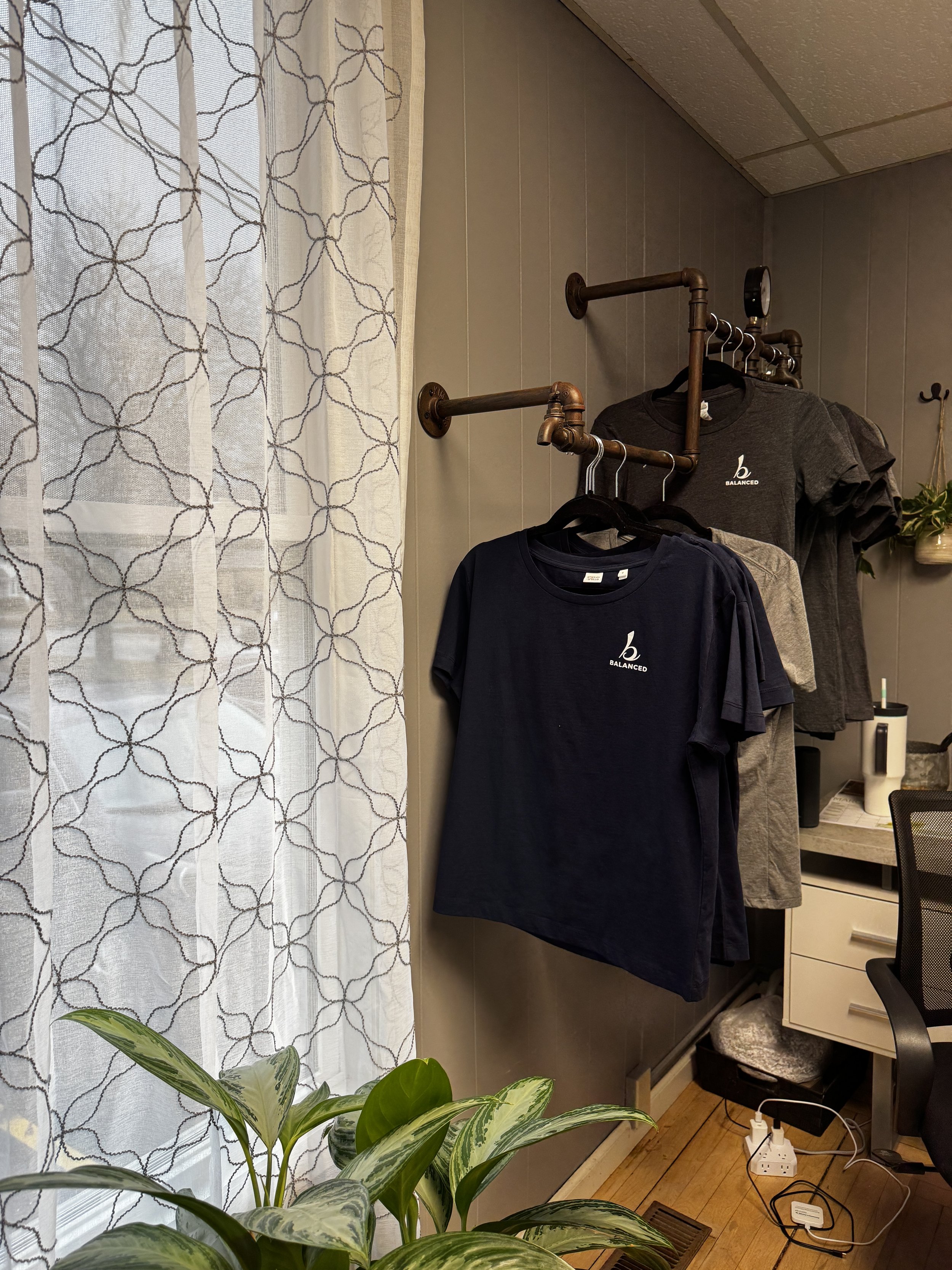 A corner of a room with a clothing rack holding black and grey T-shirts, a window with white patterned curtains, green houseplants on the floor, and a small dresser or cabinet with various items on top, including a small plant and electronic devices.