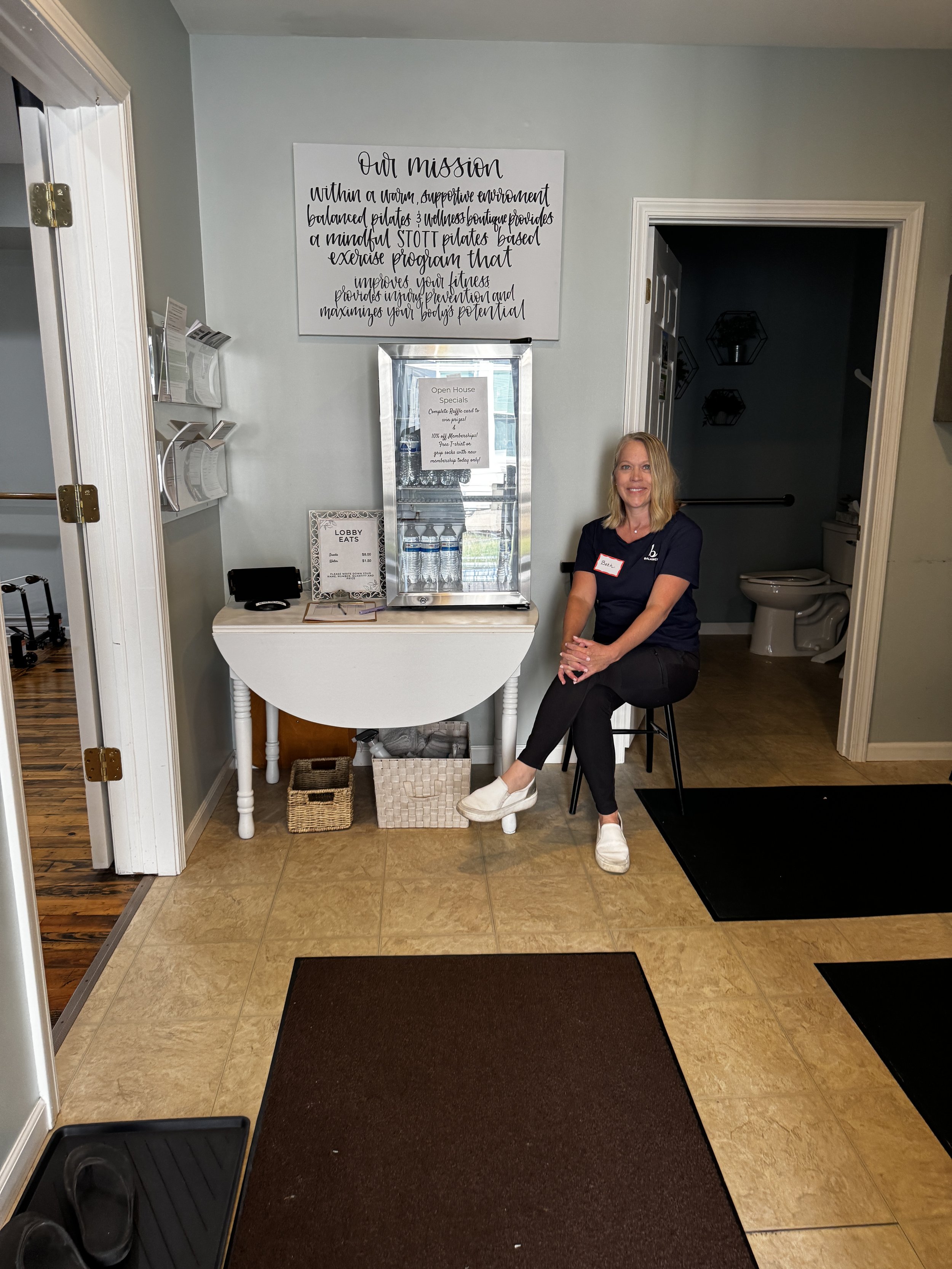 A woman sitting on a black stool at a reception area or lobby. She is wearing a navy blue shirt with a name tag and white shoes. Behind her, there is a small table with water bottles, a mirror display, and a sign. To the right, there is a bathroom wi
