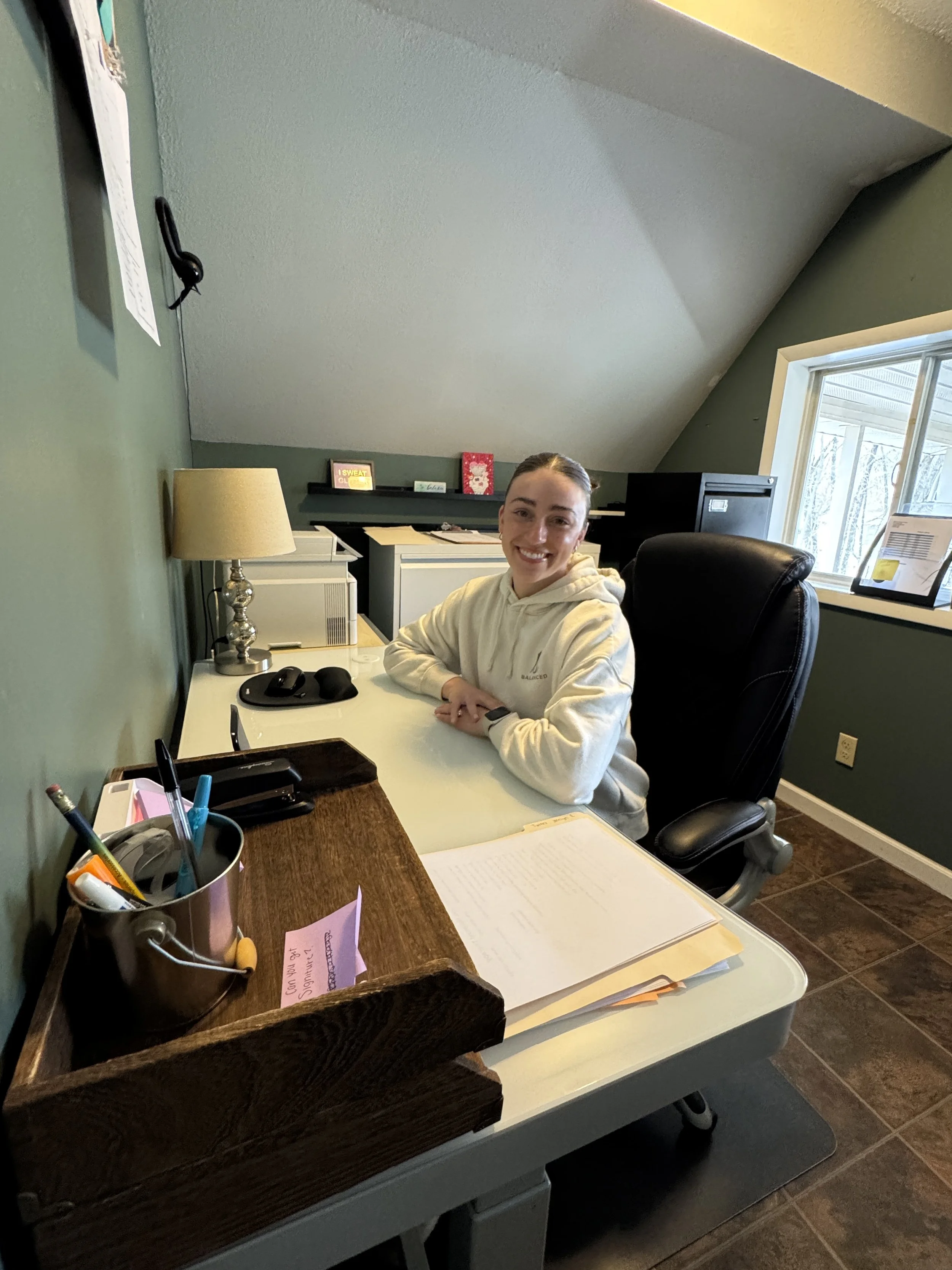 A woman sitting at a desk in an office, smiling at the camera. The desk has a lamp, a computer mouse, a pen holder, and some papers. There is a window with trees outside, and a black filing cabinet. The wall behind her has a few framed pictures and n