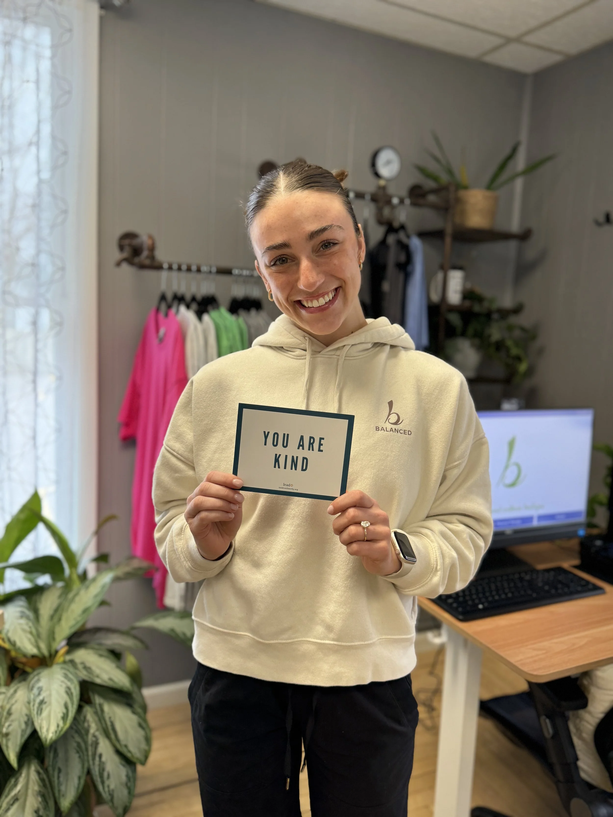Smiling woman in cream hoodie holding a sign that says "You Are Kind" inside at Balanced Pilates and Wellness Boutique in Midland, Michigan.