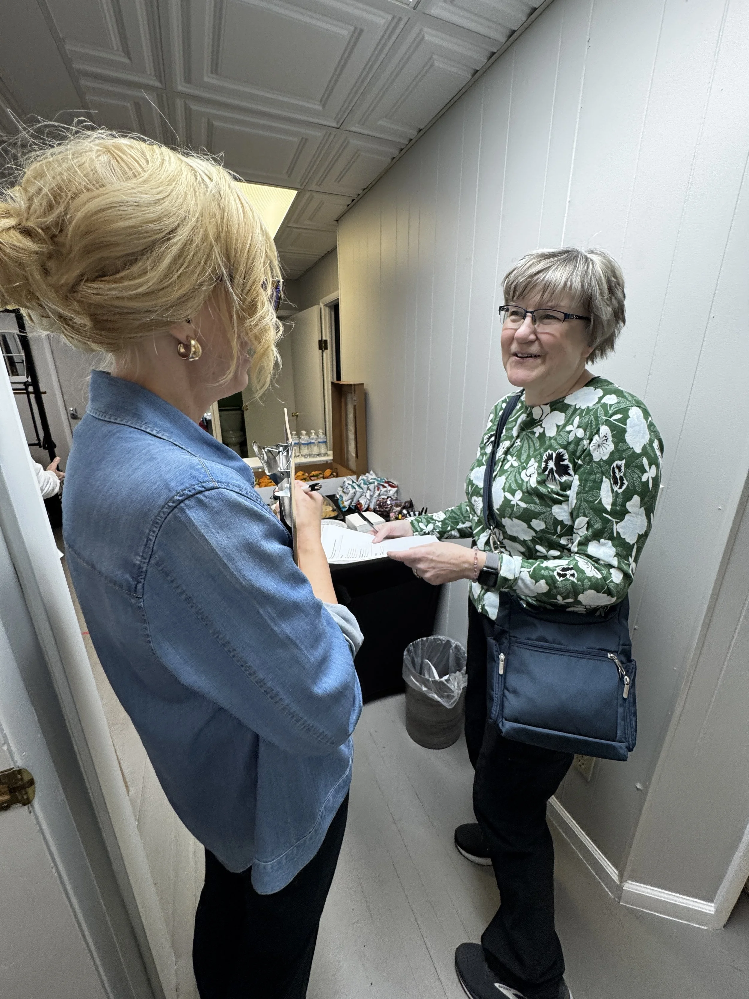 Two women are talking in a hallway, one wearing a denim jacket and the other with a green floral shirt and glasses. They are smiling and holding papers, with a table, water bottles, and snacks behind them.