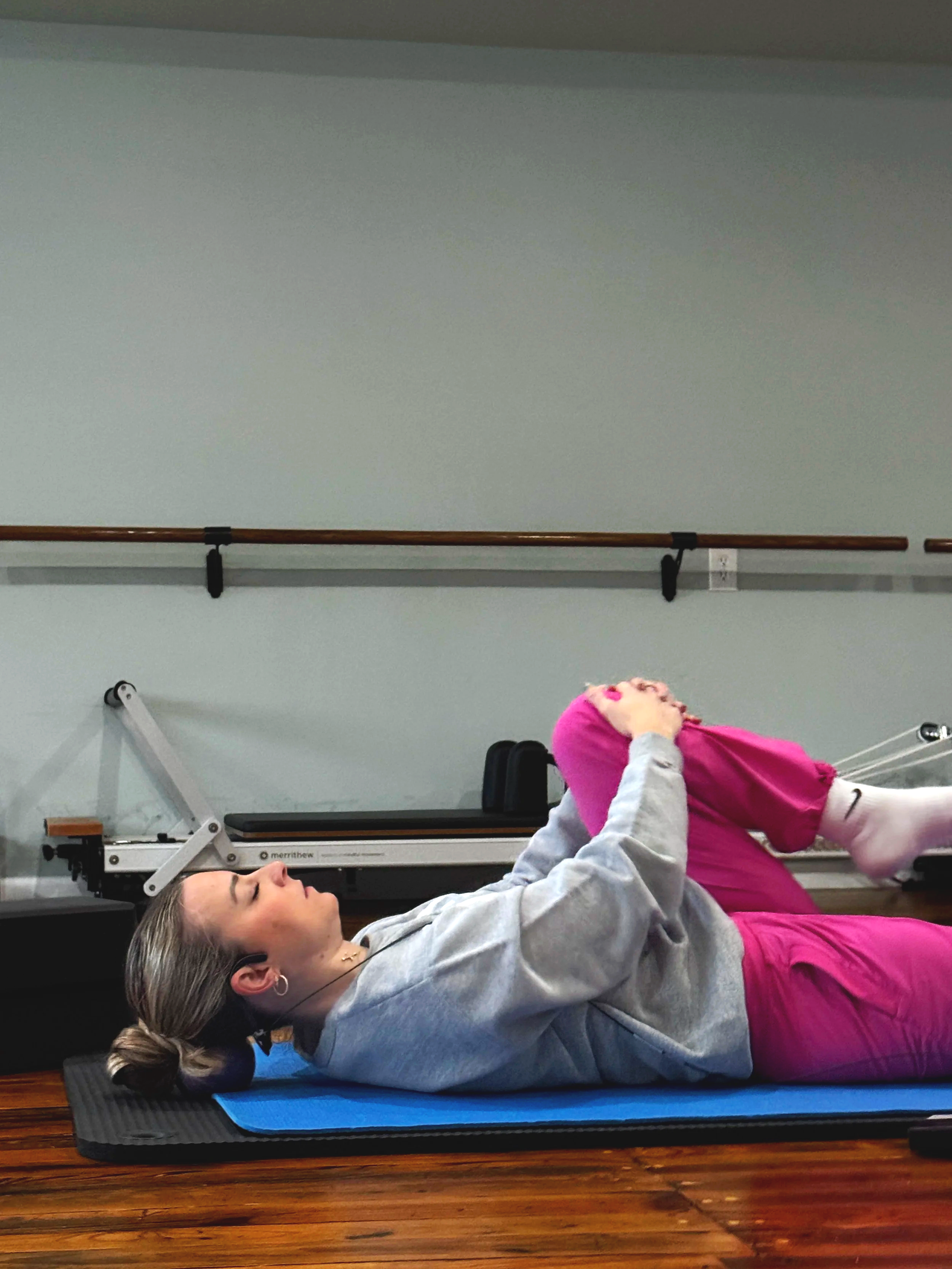 A woman lying on a Pilates mat on her back, holding her legs straight up while a young girl in pink pants and a gray sweatshirt supports her legs with her hands, in a room with wooden flooring and a ballet barre on the wall.