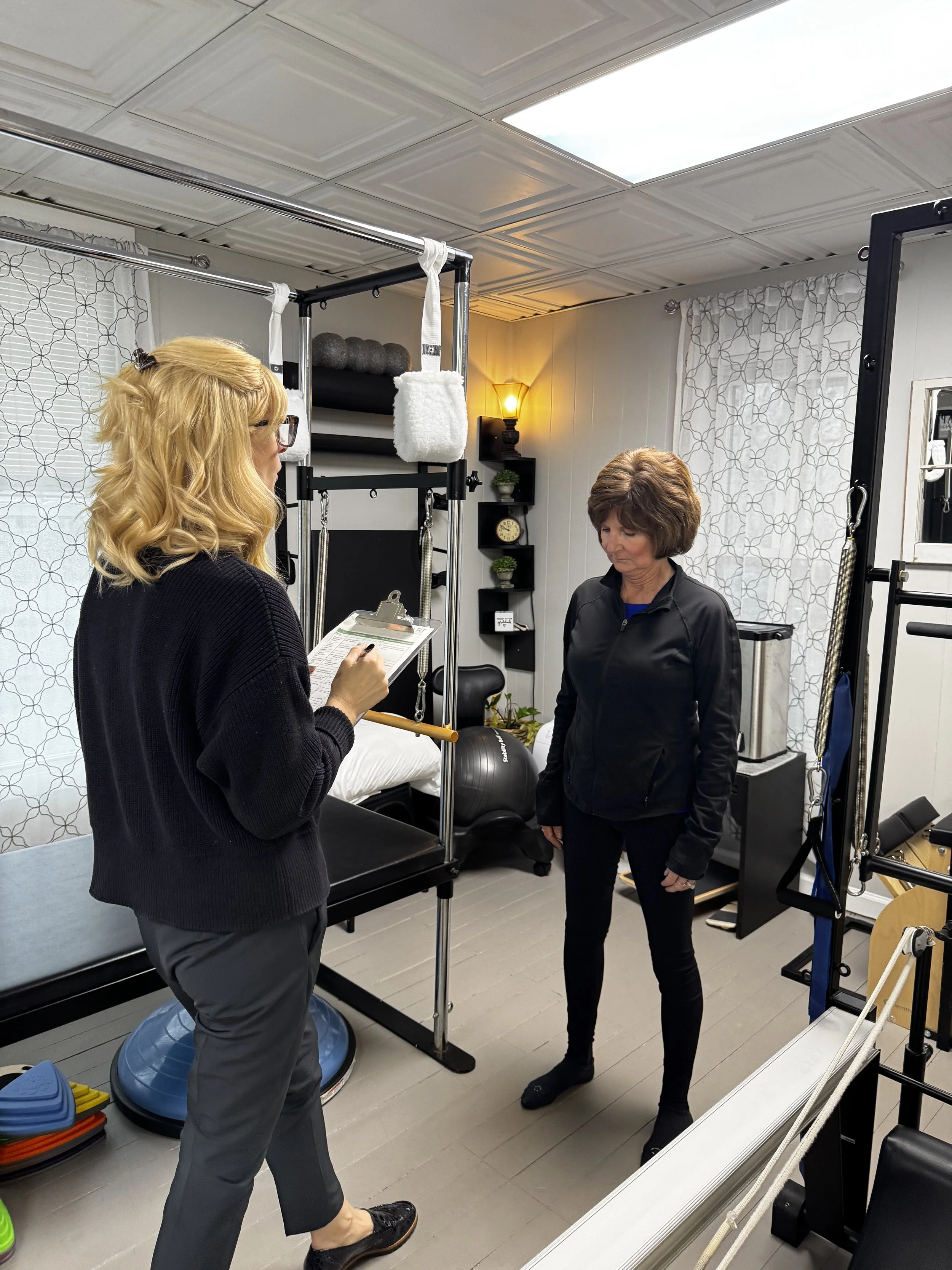 A physical therapy session in progress with two women, one holding a clipboard and appearing to give instructions to the other, in a room equipped with exercise and therapy equipment.