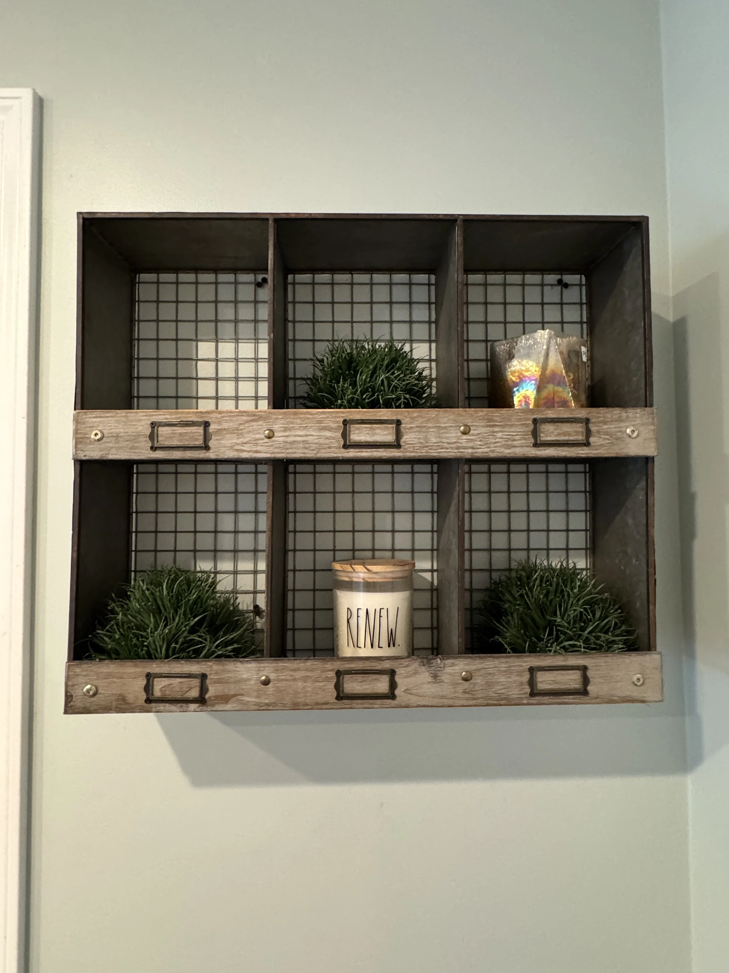 Decorative wall shelf with three sections, each holding artificial greenery, a candle labeled "RENEW," and a crystal or glass ornament, mounted on a light green wall.