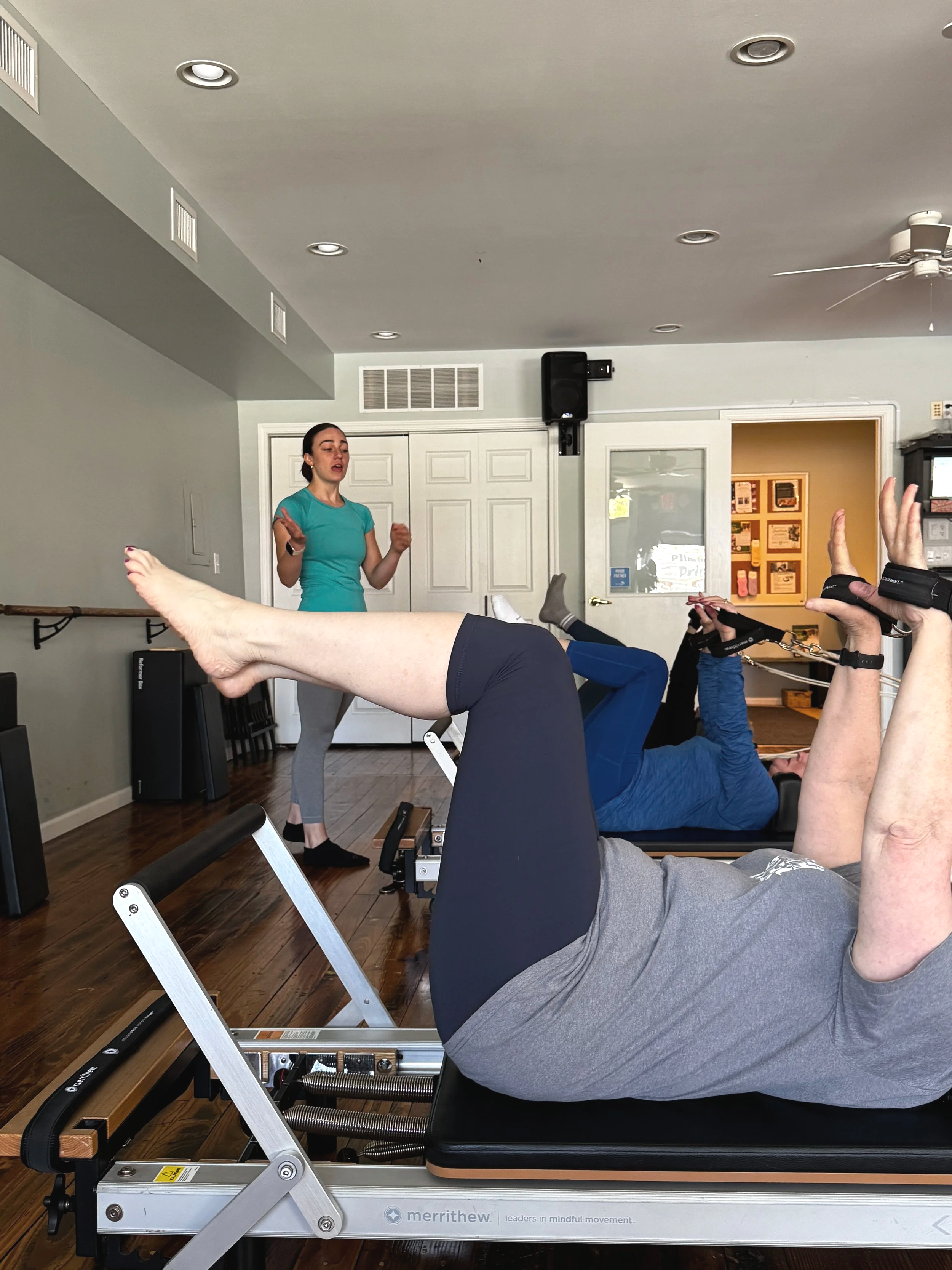 A woman giving instructions to a person lying on a Pilates reformer machine in a fitness studio.