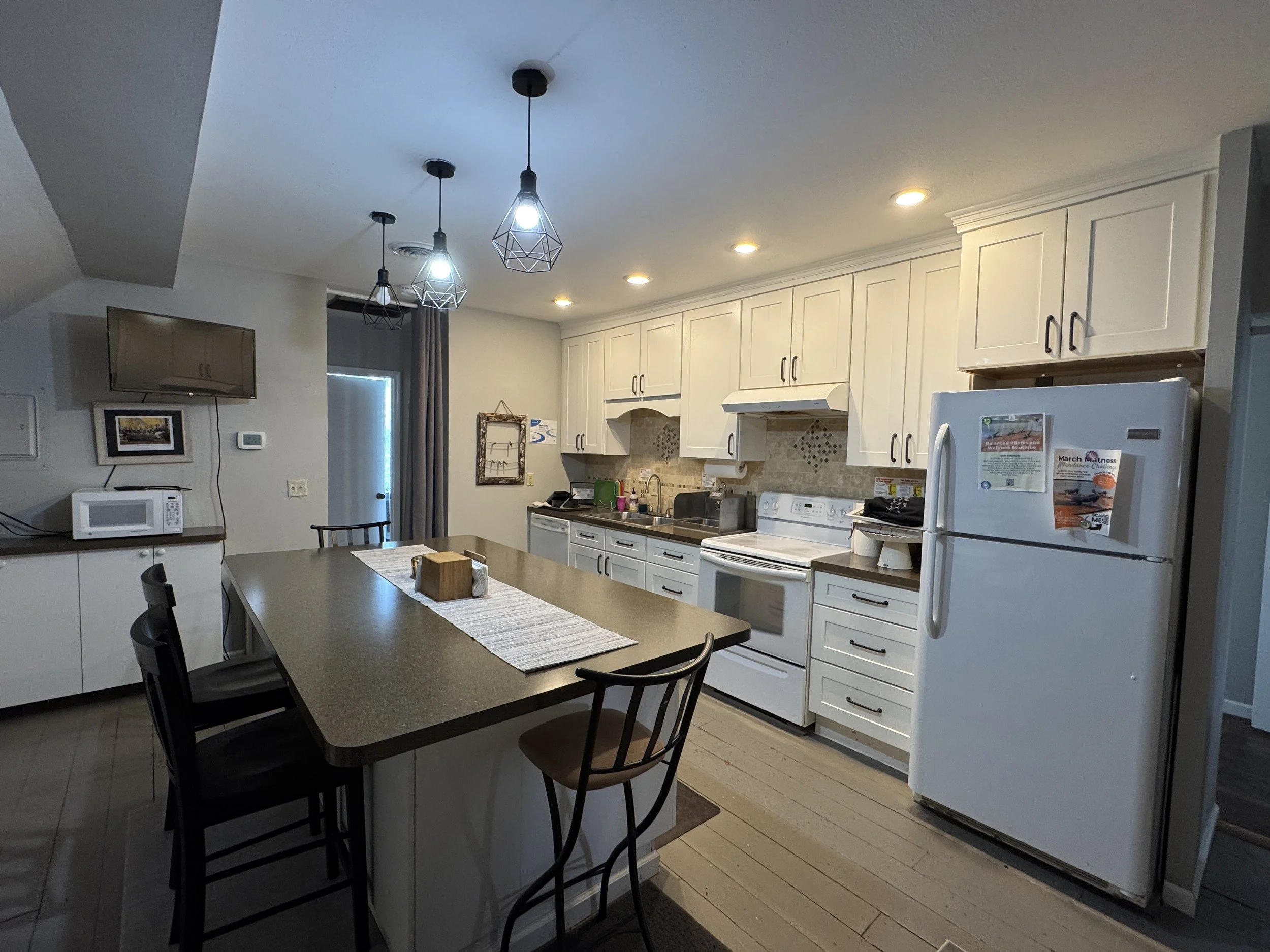 Kitchen with white cabinets and black hardware, microwave, washing machine, and refrigerator, pendant lights, window with curtain, and dining table with chairs.