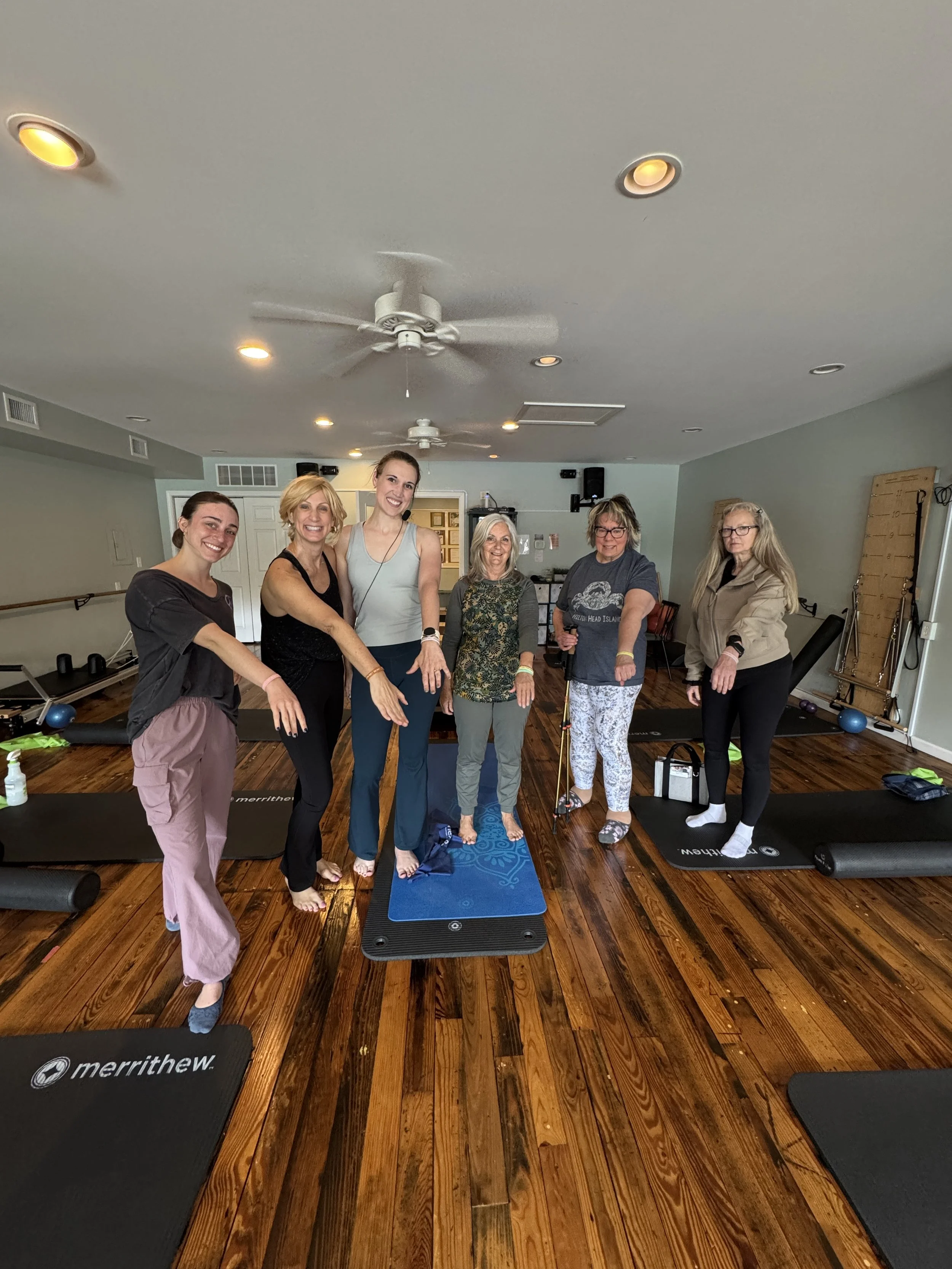 Group of six women standing together in a Pilates studio, smiling and posing for a photo, with exercise mats and equipment on the floor.