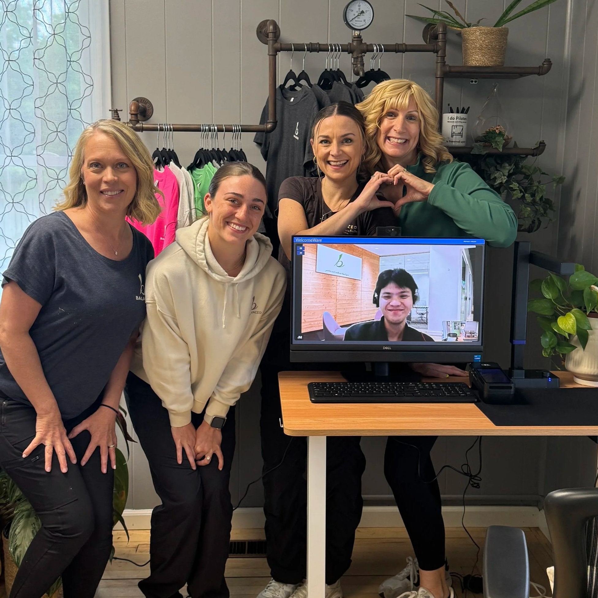 Four women smiling in front of a computer and clothing rack, making a heart shape with their hands in a room with plants and a window at Balanced Pilates and Wellness Boutique in Midland, Michigan.
