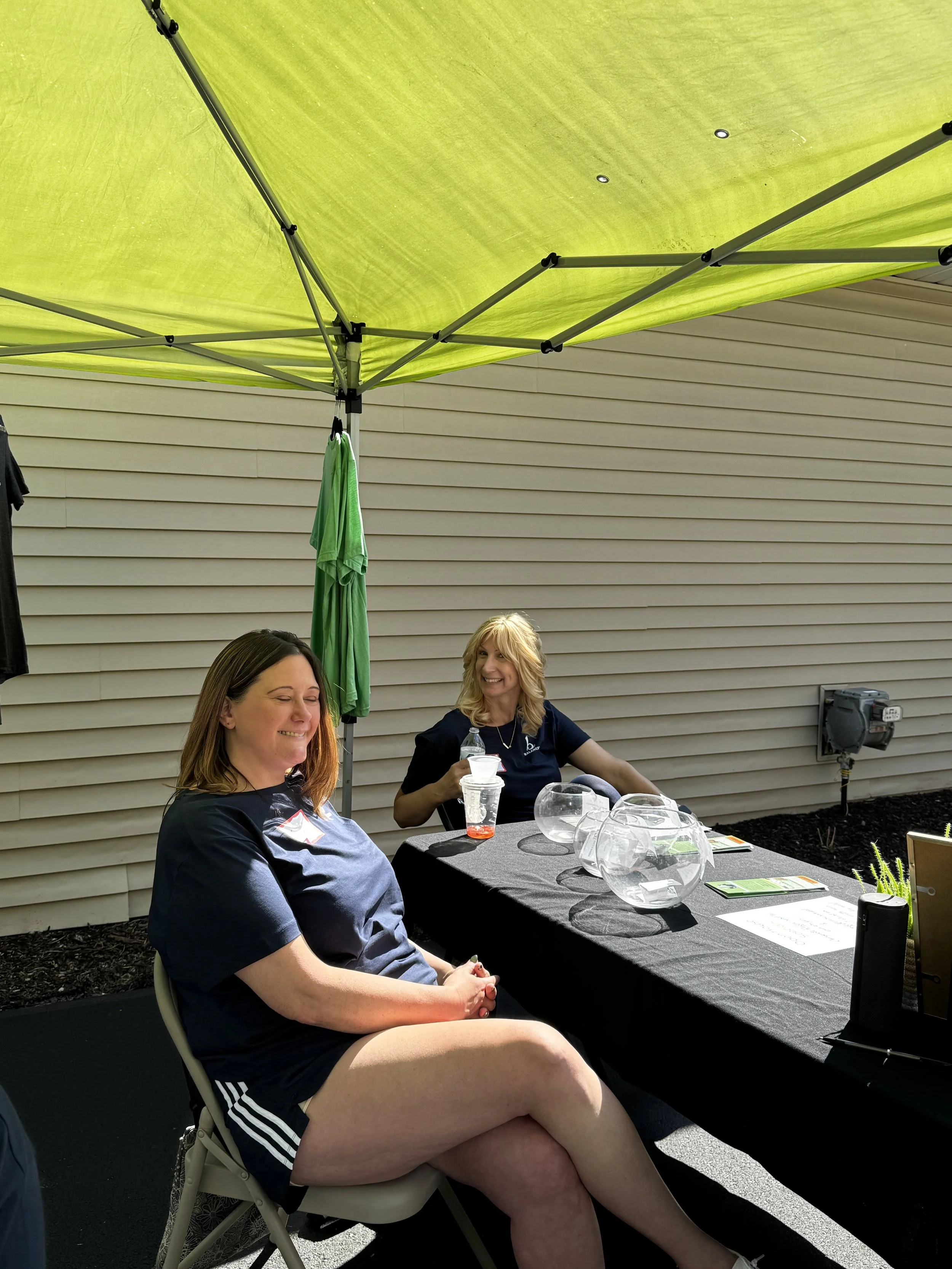 Two women sitting at a table outdoors, smiling, with a green canopy overhead, in front of a beige house with horizontal siding. The table is covered with a black tablecloth and has clear glass bowls, a water bottle, and some papers on it.