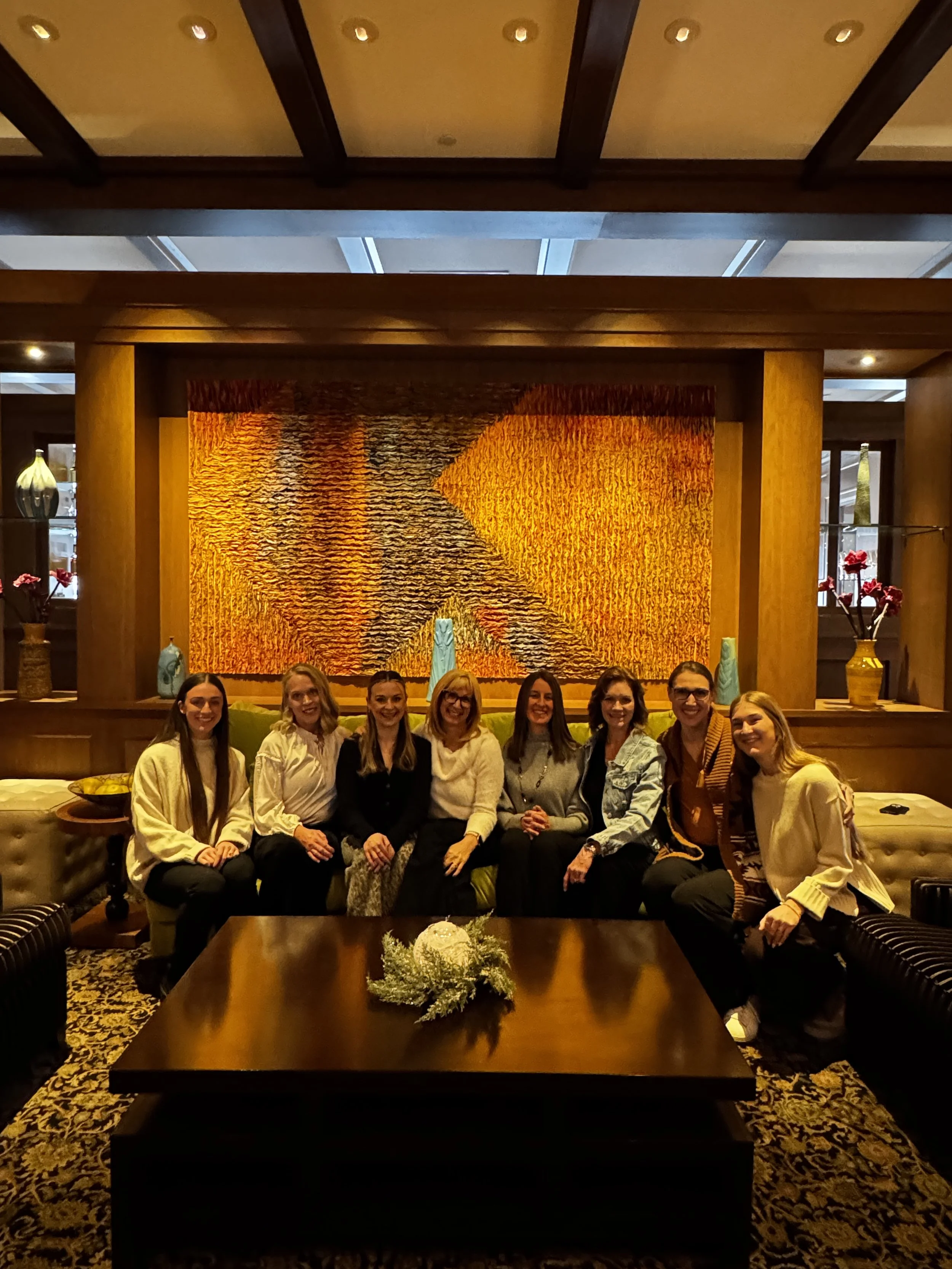 A group of eight women sitting on a green couch in front of a large orange and brown textured art piece in a warmly lit room. The women are smiling and dressed in casual to semi-formal attire, with a coffee table in front of them decorated with a whi