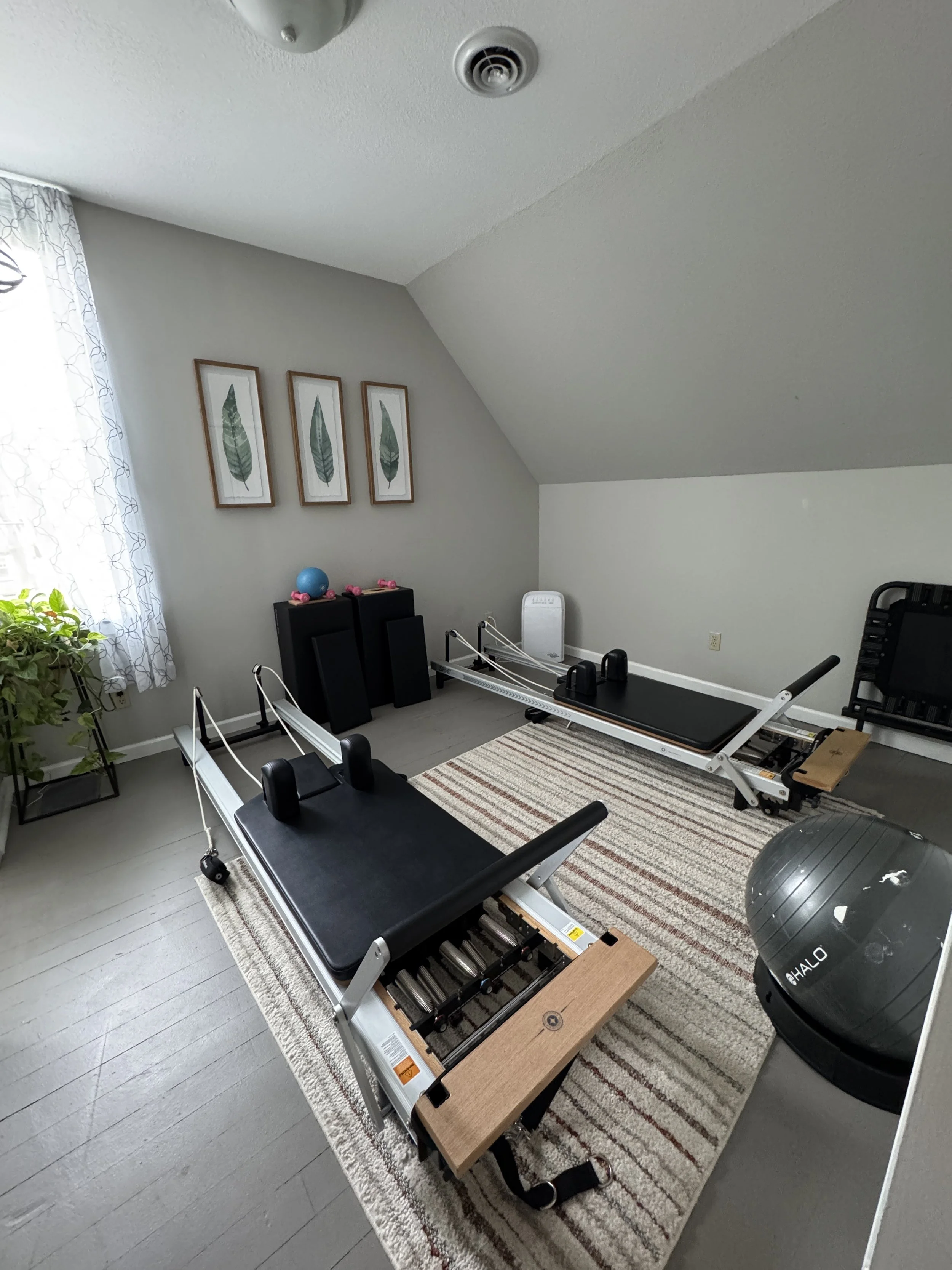 Room with a reformer Pilates machine, exercise ball, and fitness equipment on a striped rug, with framed leaf artwork on the wall and a window with sheer curtains.
