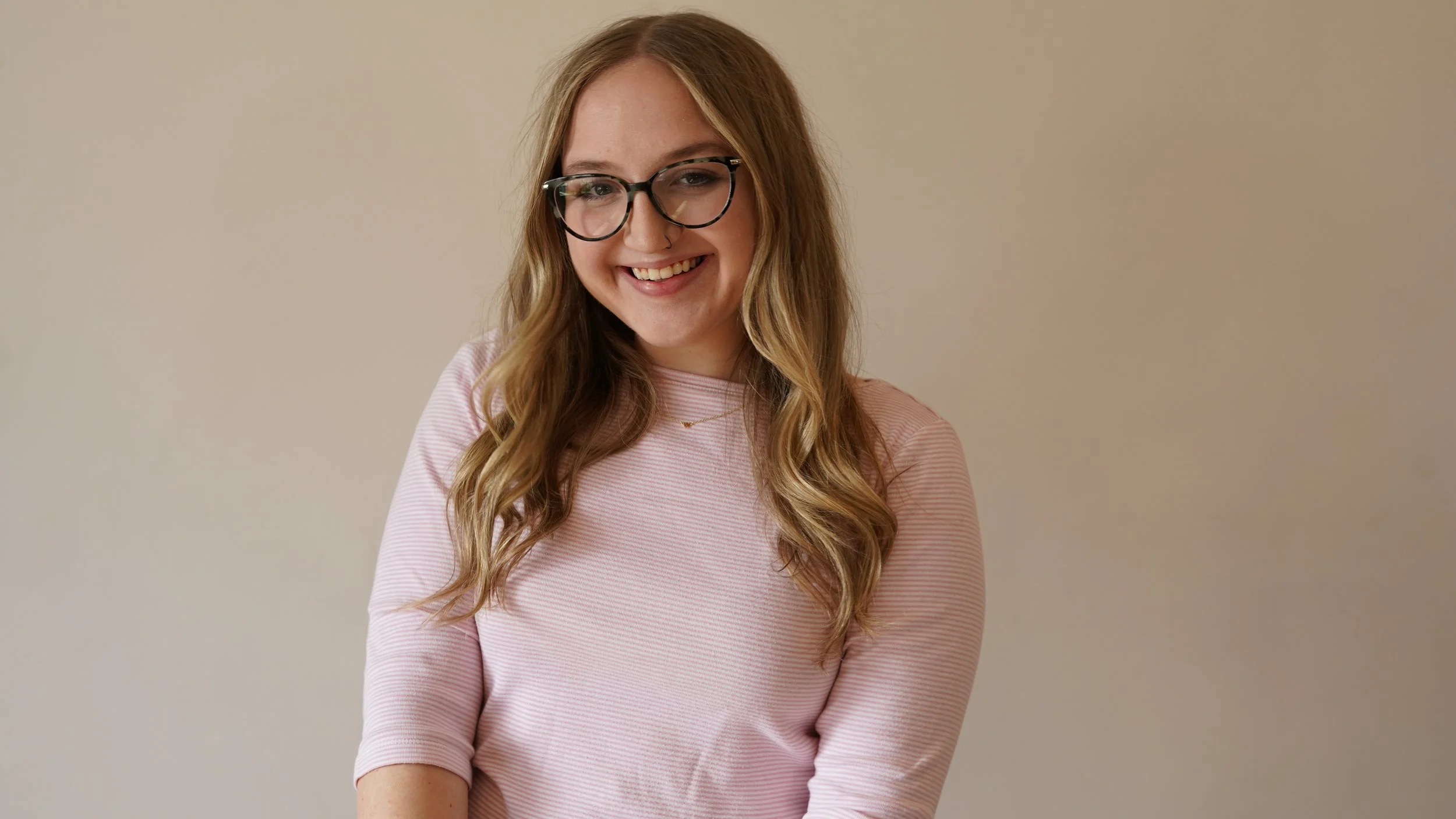 Mikayla Lewis, marketing assistant, wearing large black glasses and a pink and white striped shirt, standing against a plain beige wall.