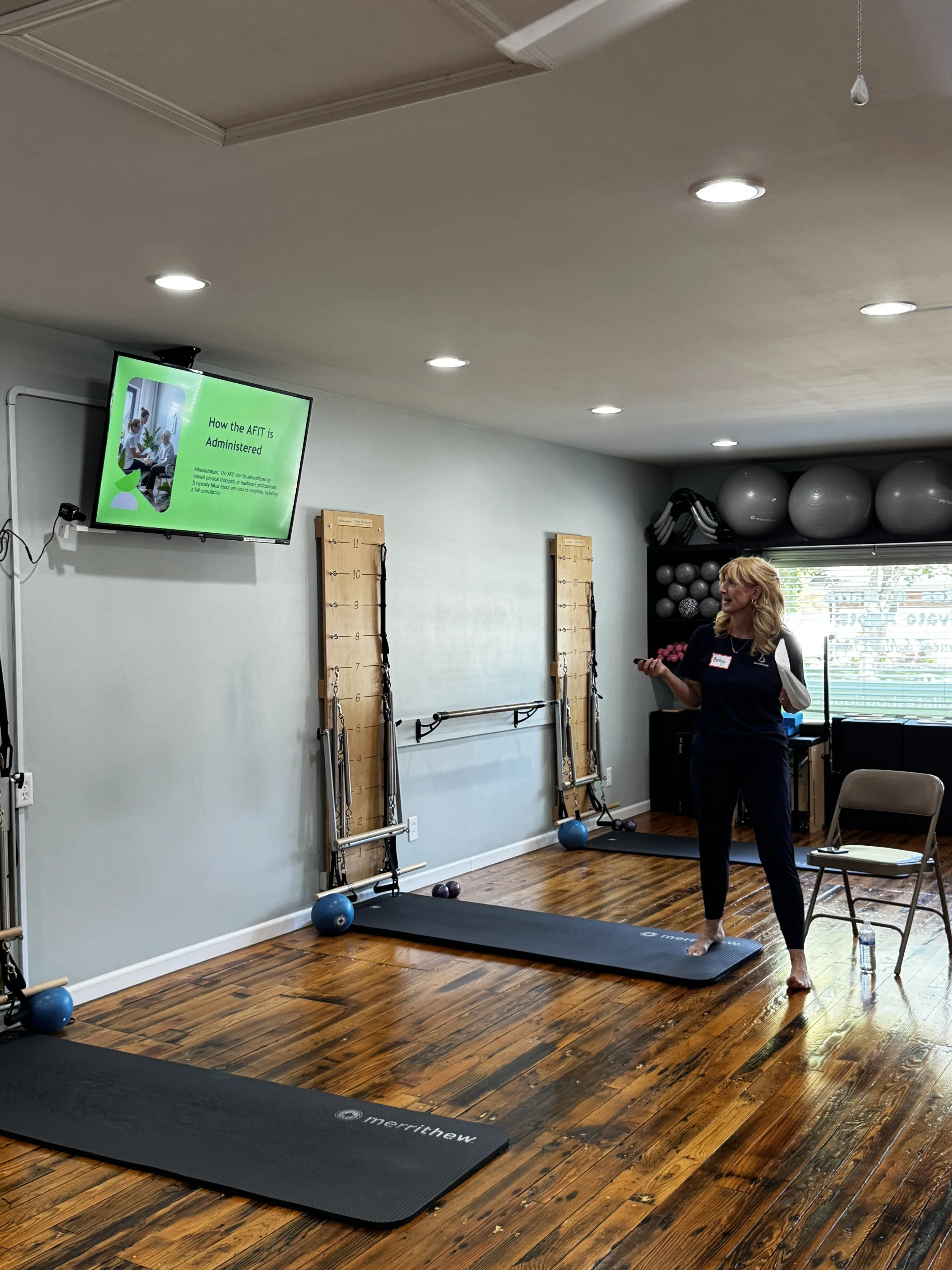 A woman in workout attire standing on a yoga mat in a Pilates studio with wooden floors, exercise balls on a shelf, and a screen displaying a presentation titled 'How the AFIT is Administered'.