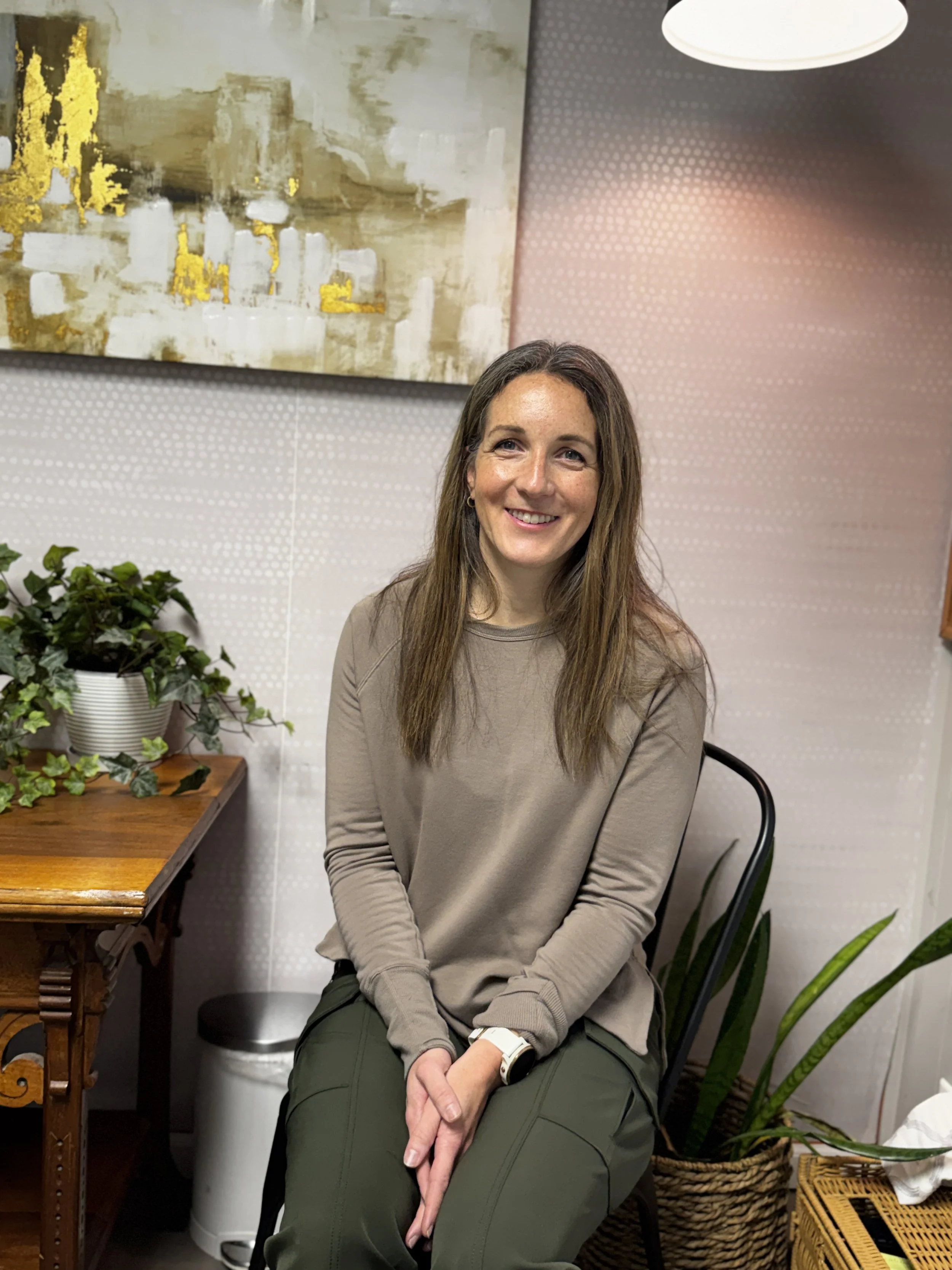 A woman with brown hair and a beige long-sleeve top sitting on a black chair indoors with a wall decorated with abstract art and plants around her.