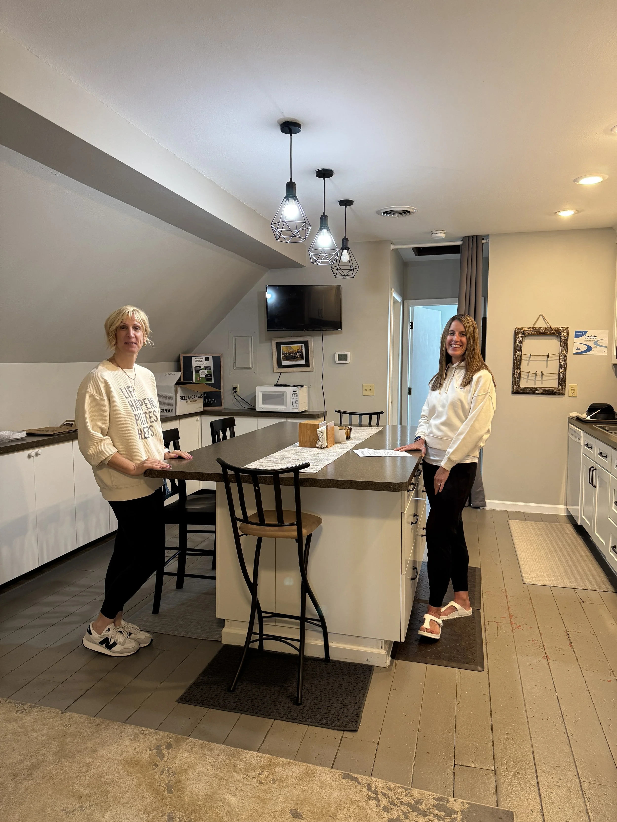 Two women standing in a kitchen, smiling at the camera. One woman has blonde hair and is wearing a cream-colored sweatshirt and sneakers, the other has brown hair and is wearing a white hoodie and sandals. The kitchen has white cabinets, a microwave,