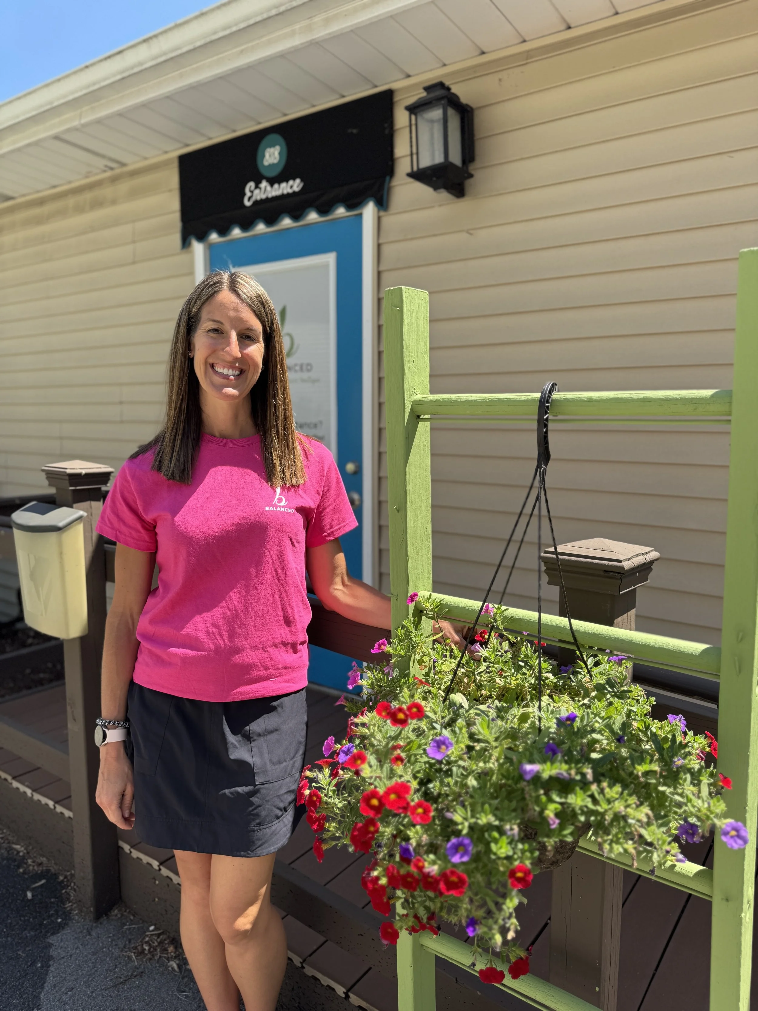 A smiling woman in a pink shirt standing next to colorful hanging flowers outside a building with beige siding, a blue door, and a black sign that reads "Entrance."