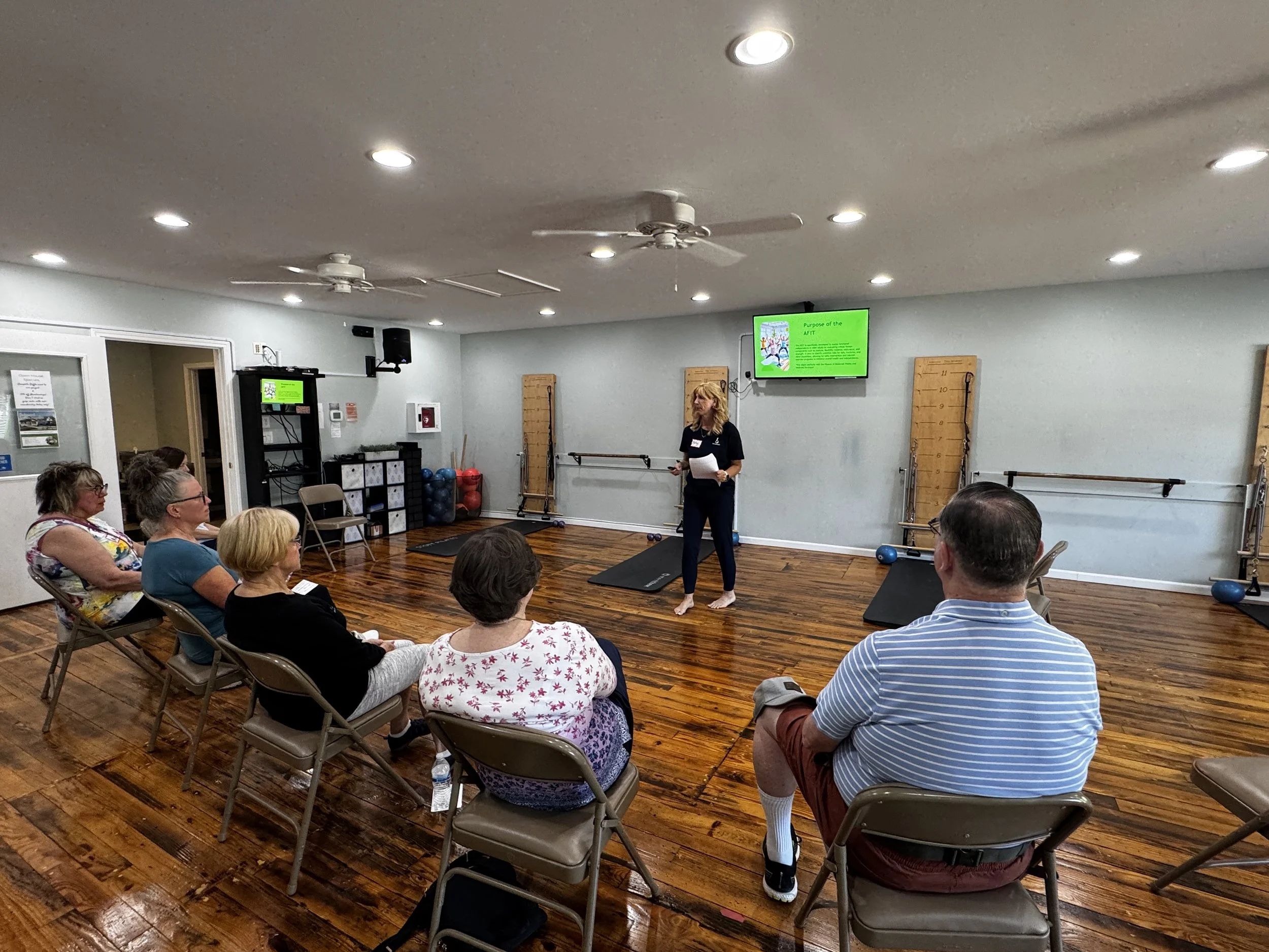 A group of people attending a presentation in a room with wooden floors, chairs, and a large screen displaying a slide titled 'Purpose of the AFIT'.