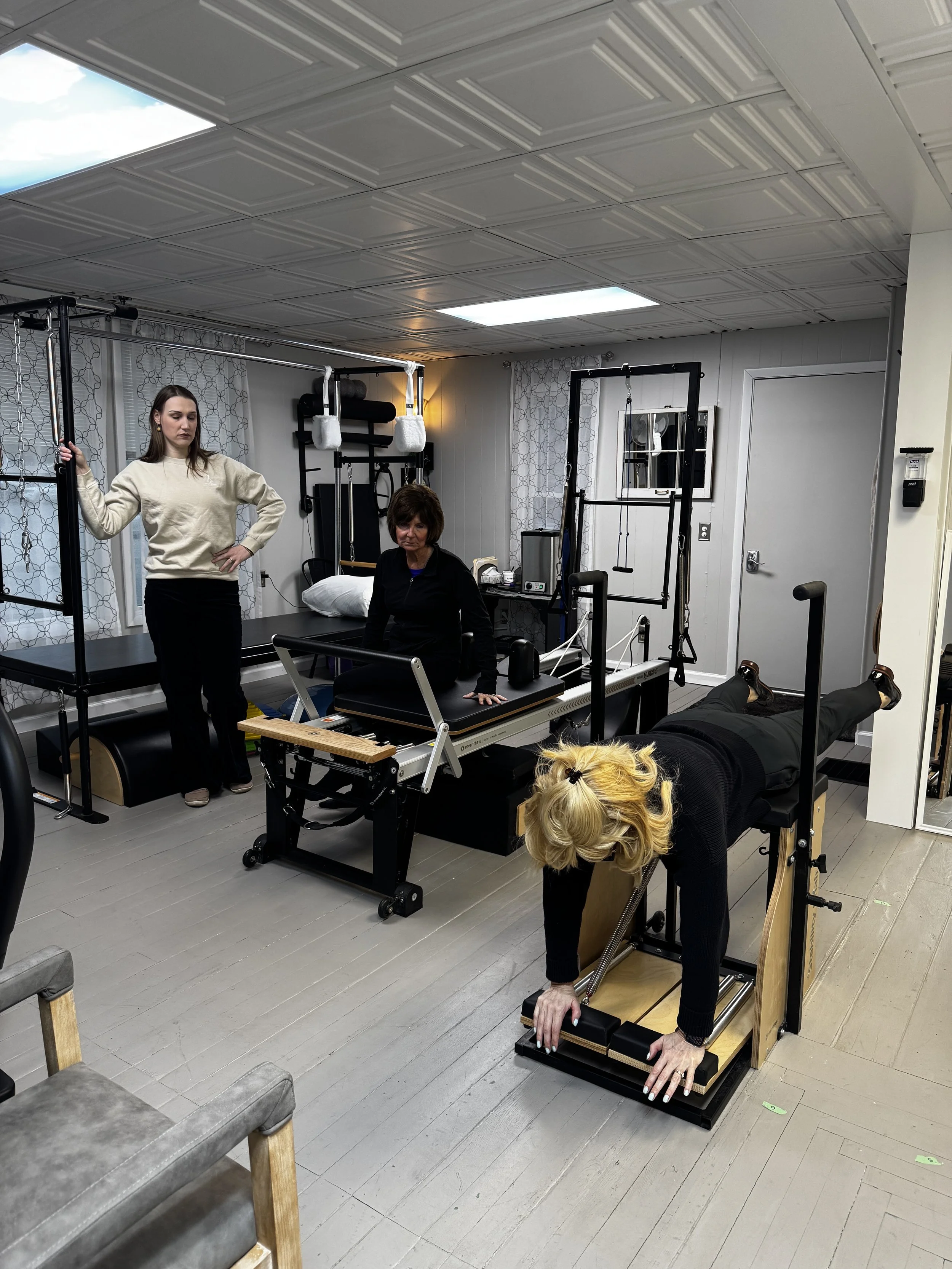 A woman is lying face down on a wooden Pilates reformer machine with her feet on the foot bar. She is holding onto the side handles of the machine while another woman, standing nearby, supervises. The room appears to be a Pilates or physical therapy 