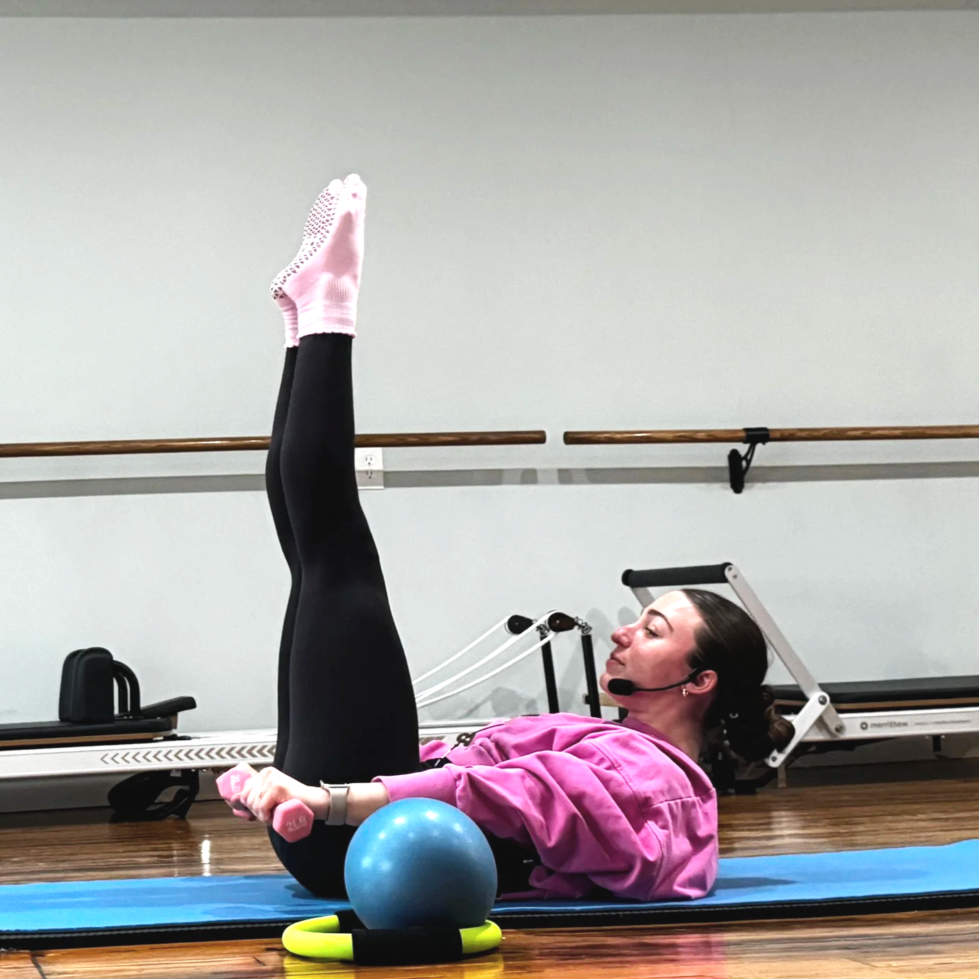 A woman exercising on a Pilates mat in a studio in Midland, Michigan, lifting her legs vertically in the air while lying on her back, with fitness equipment and a ballet barre in the background.