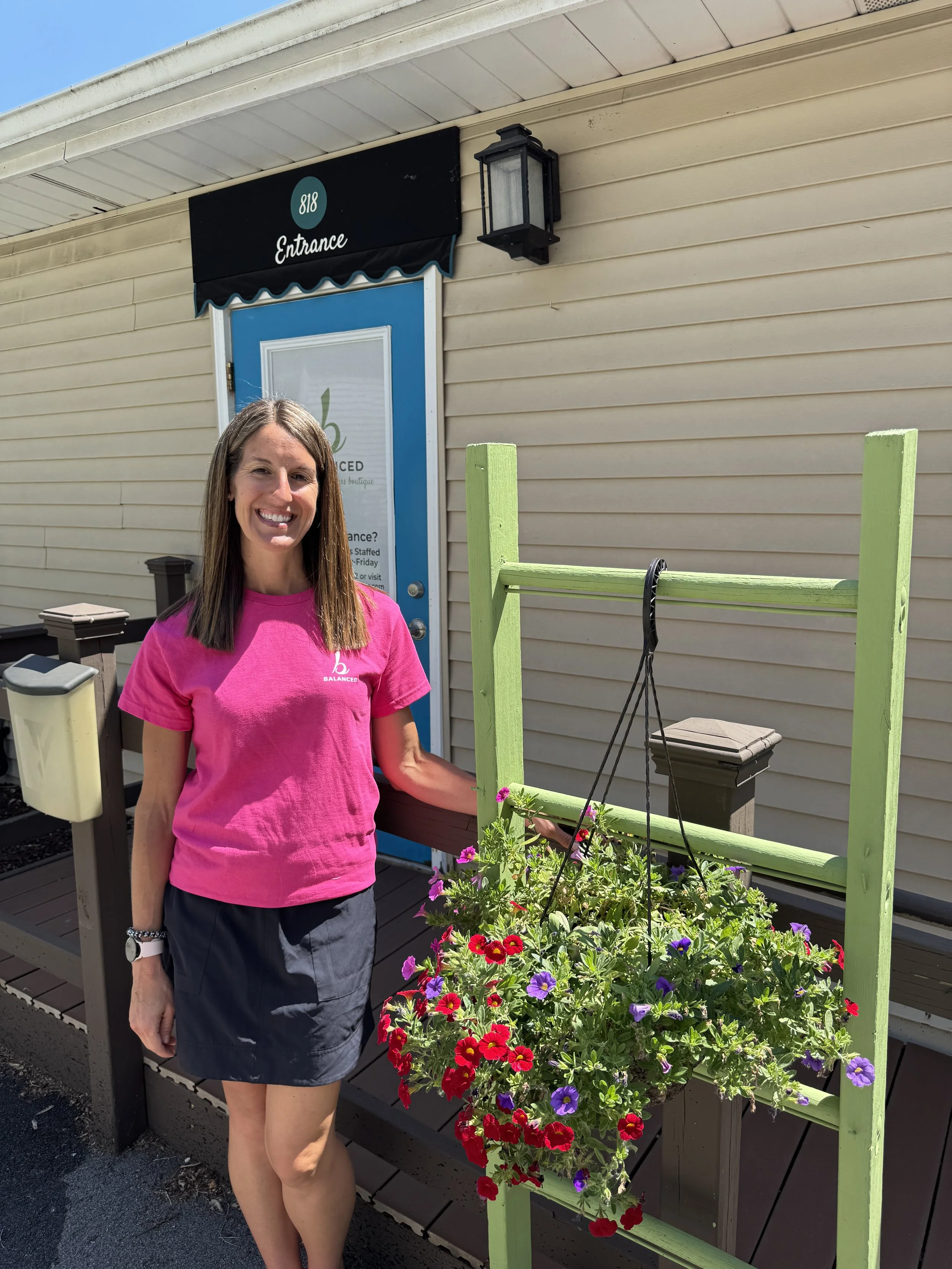 A woman standing beside a green planter filled with colorful flowers outside a building, with a blue door and beige siding, sunny weather, and a black lantern above.