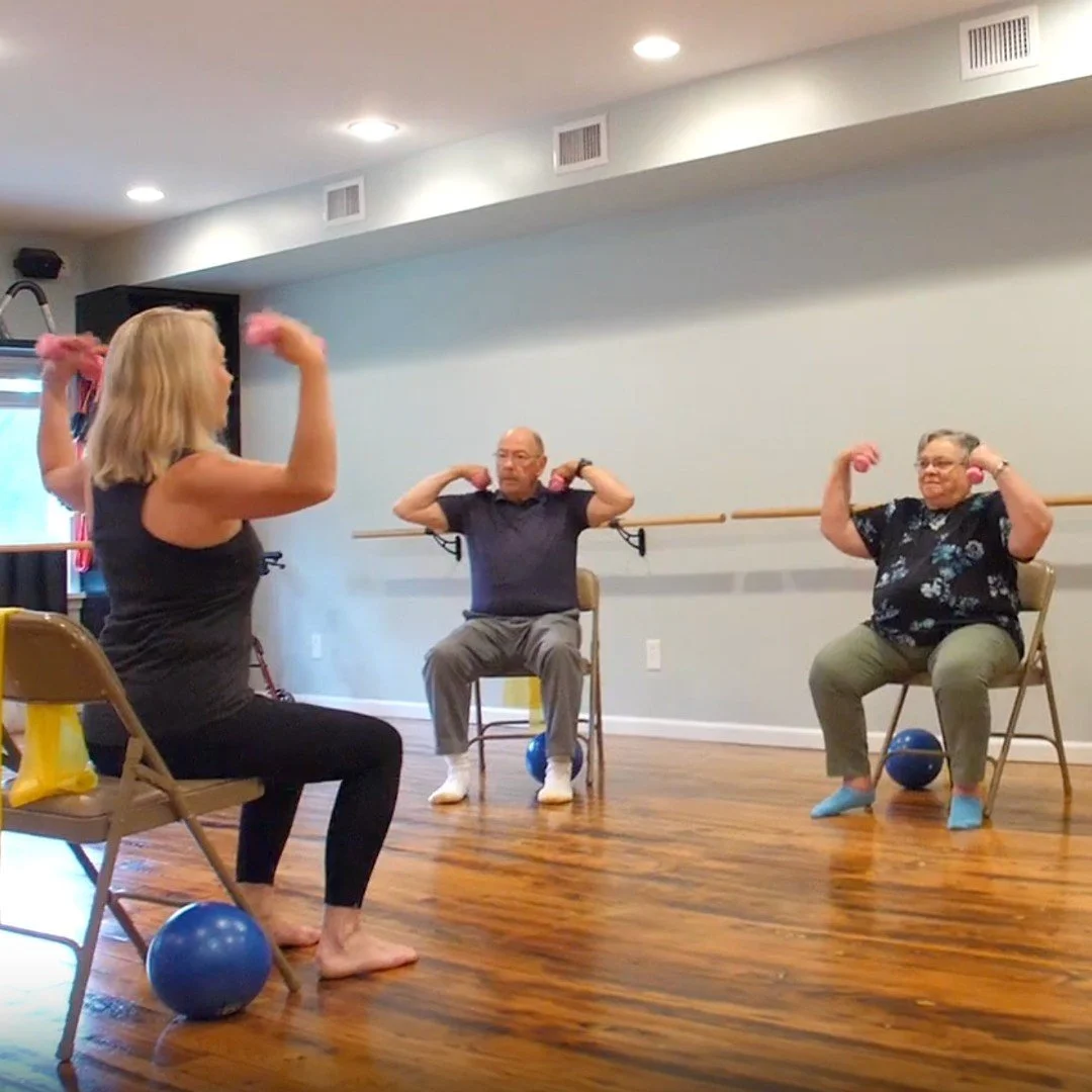 A Chair Pilates exercise class featuring senior adults seated on chairs, lifting small pink dumbbells with some blue exercise balls on the floor, in a room with hardwood floors and a ballet barre on the wall.