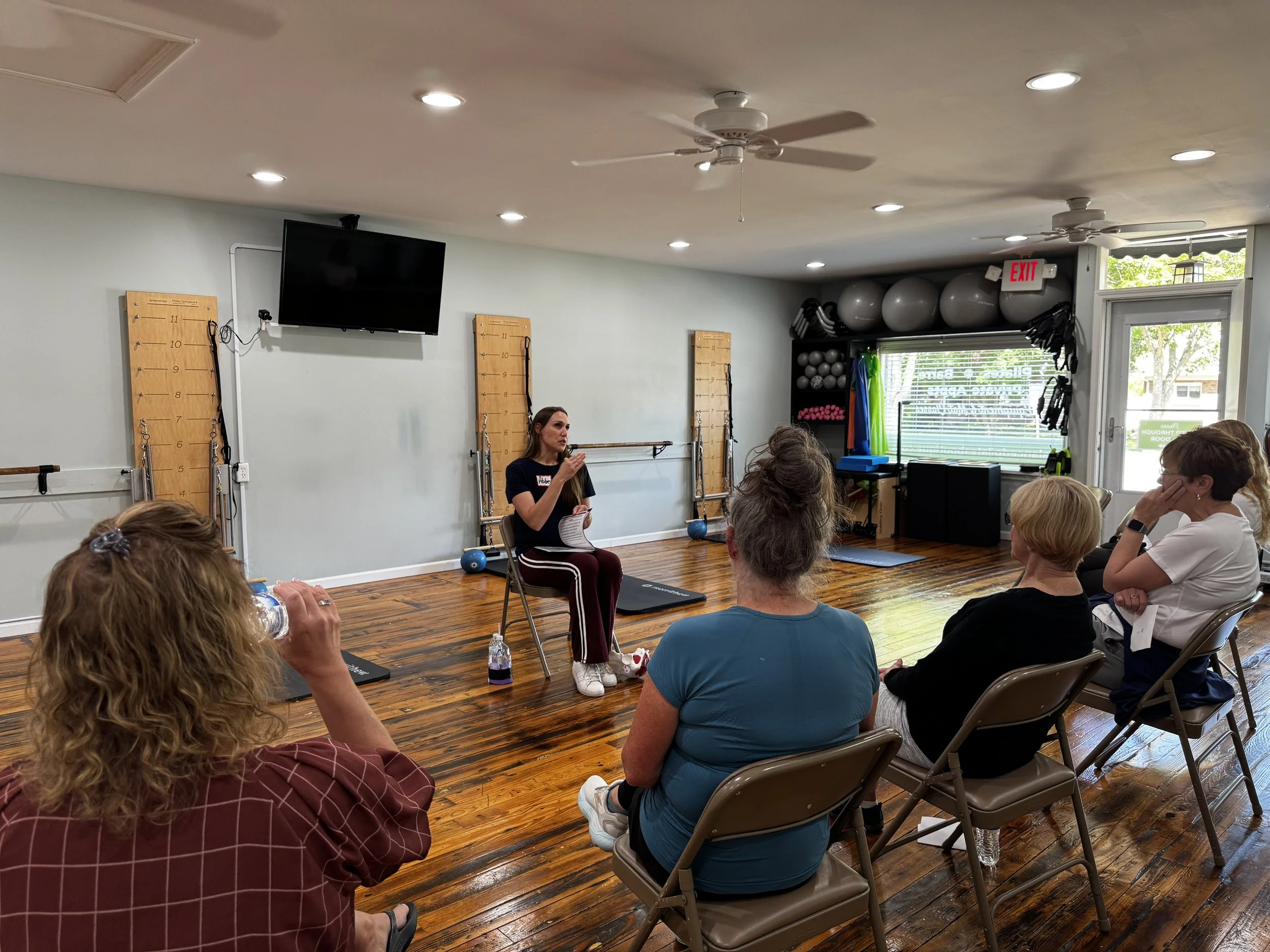 A woman sitting on a chair in front of an audience, speaking and gesturing with her hand during an education workshop in a room with exercise equipment and large windows at Balanced Pilates and Wellness Boutique in Midland, Michigan.