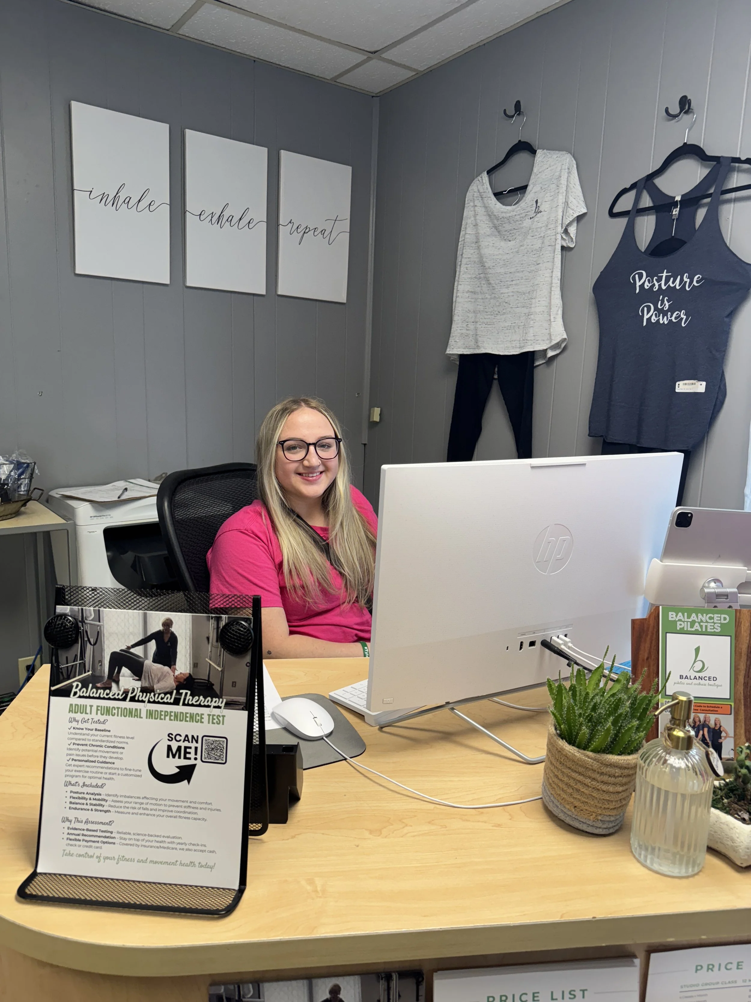 A smiling woman with long blonde hair and glasses sitting at an office desk, surrounded by various items including a white computer monitor, a potted cactus, a glass bottle, and a sign promoting balanced physical therapy and adult functional independ
