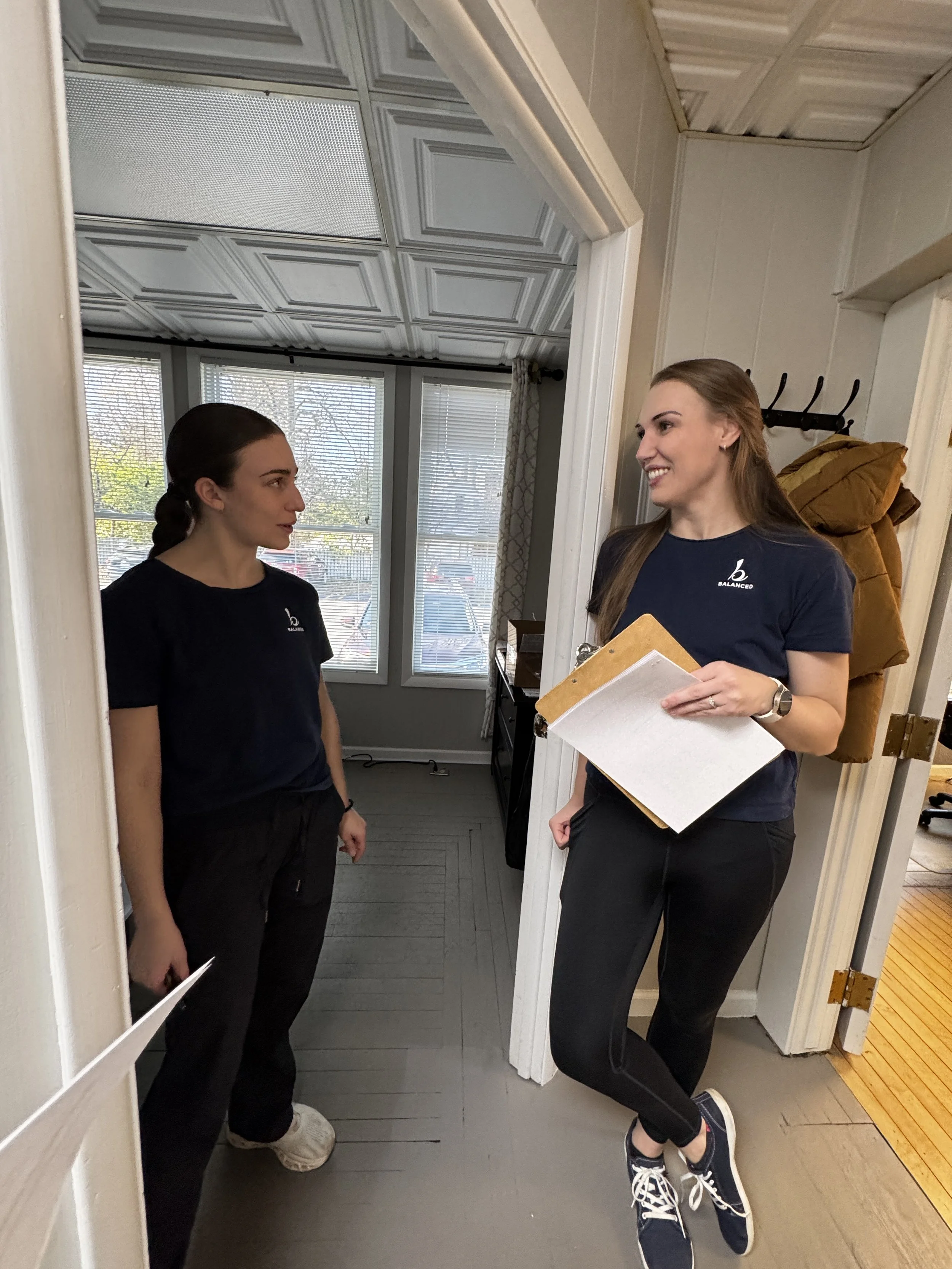 Two women are standing and talking indoors, one is facing the mirror and the other is facing away, holding wood-like sheets with papers on top.