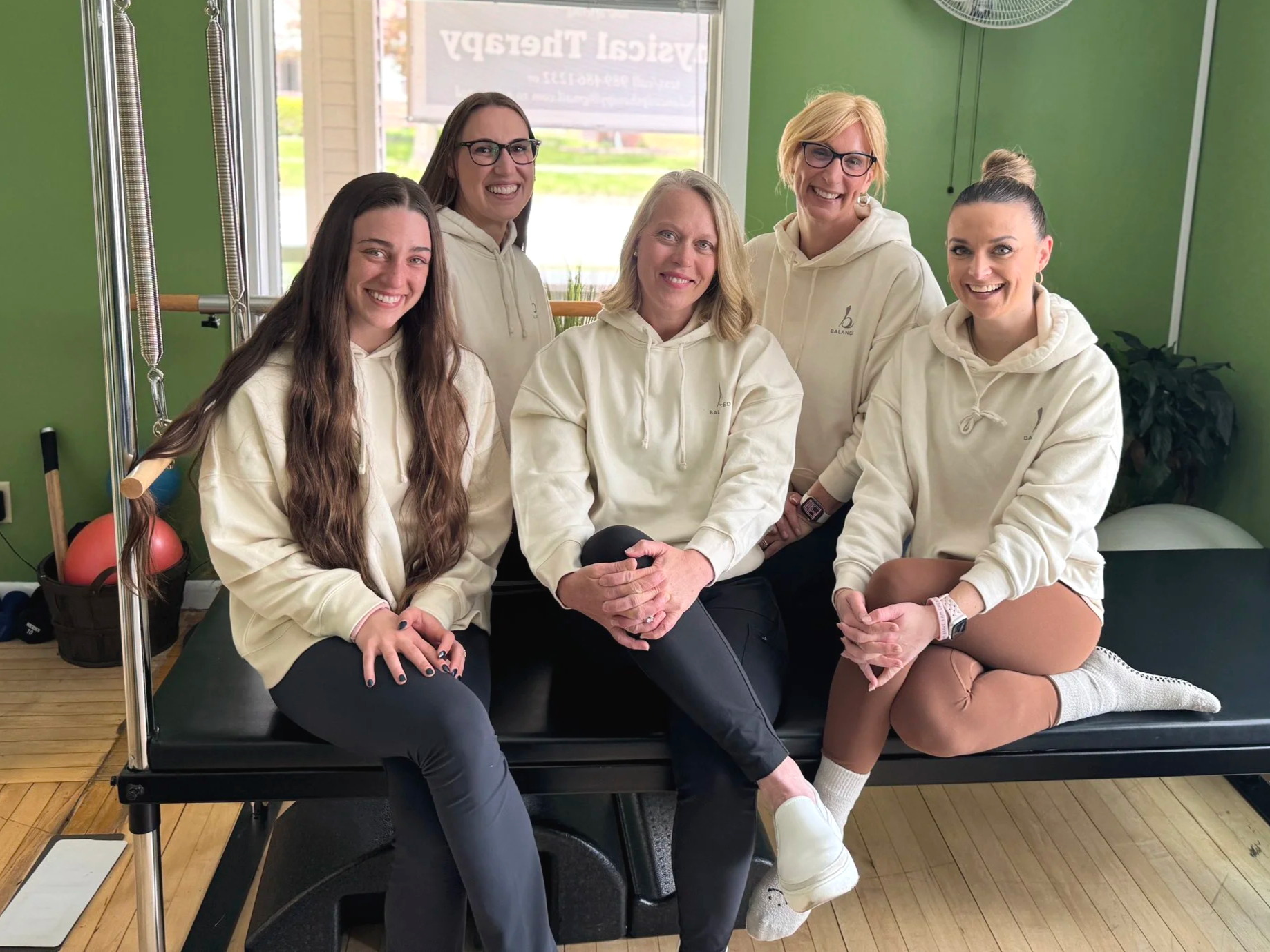 Women sitting on a black workout bench inside a gym with green walls, smiling at the camera. They are wearing beige hoodies with a logo, some with glasses. The background includes gym equipment and a window with a sign reading 'Physical Therapy'.