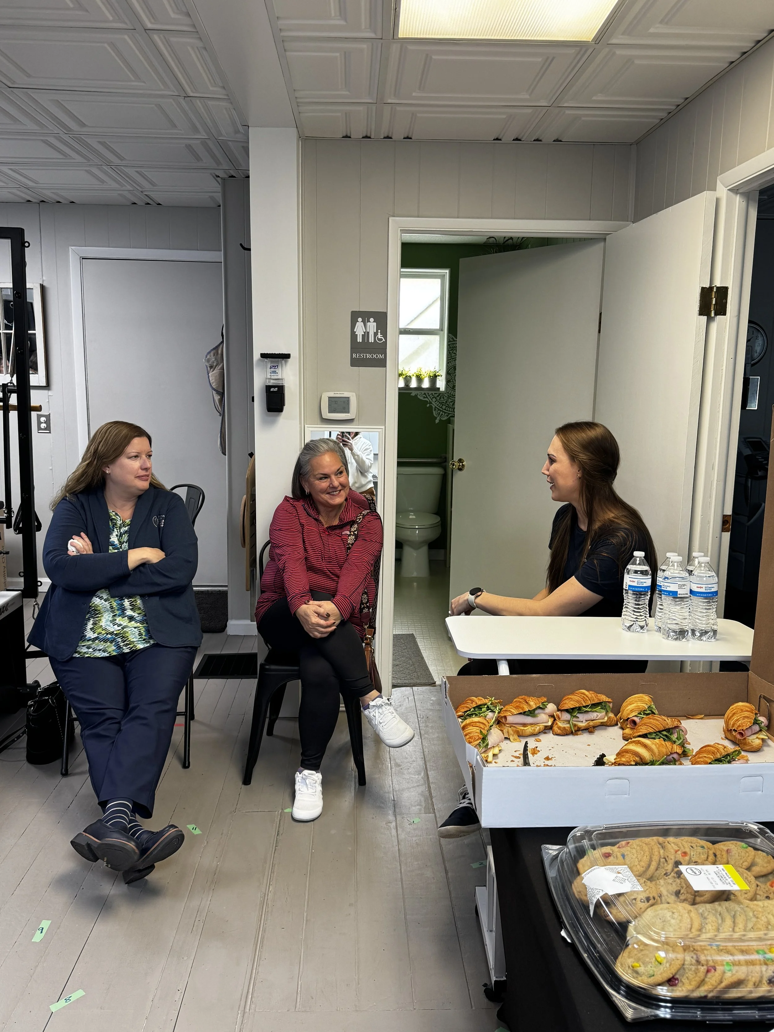 People sitting at a table with trays of sandwiches, cookies, and bottled water in a room with visible restroom signs on the wall.