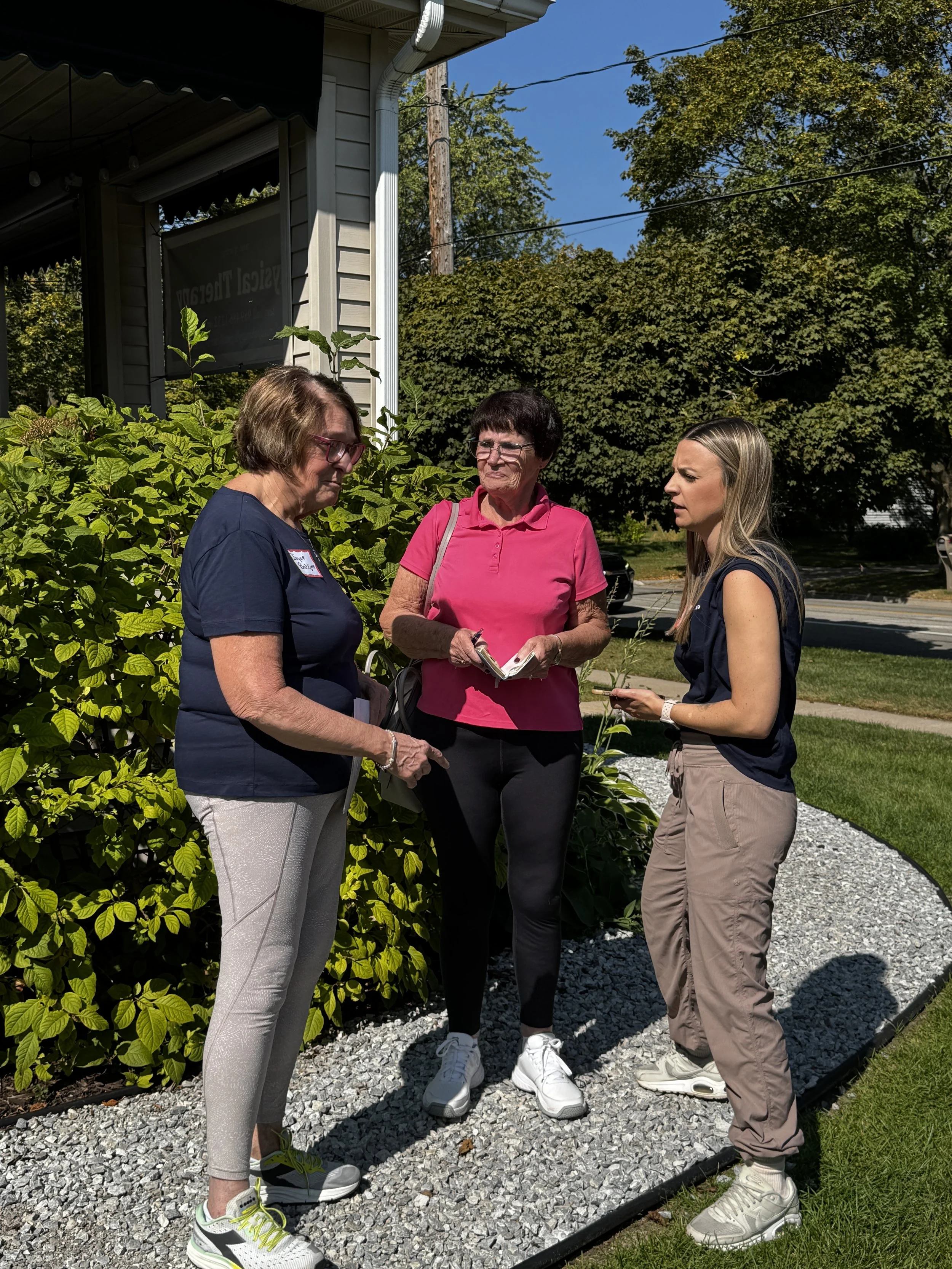 Three women having a conversation outdoors near at Balanced Pilates and Wellness Boutique in Midland, Michigan with trees in the background, two are holding notebooks or papers.