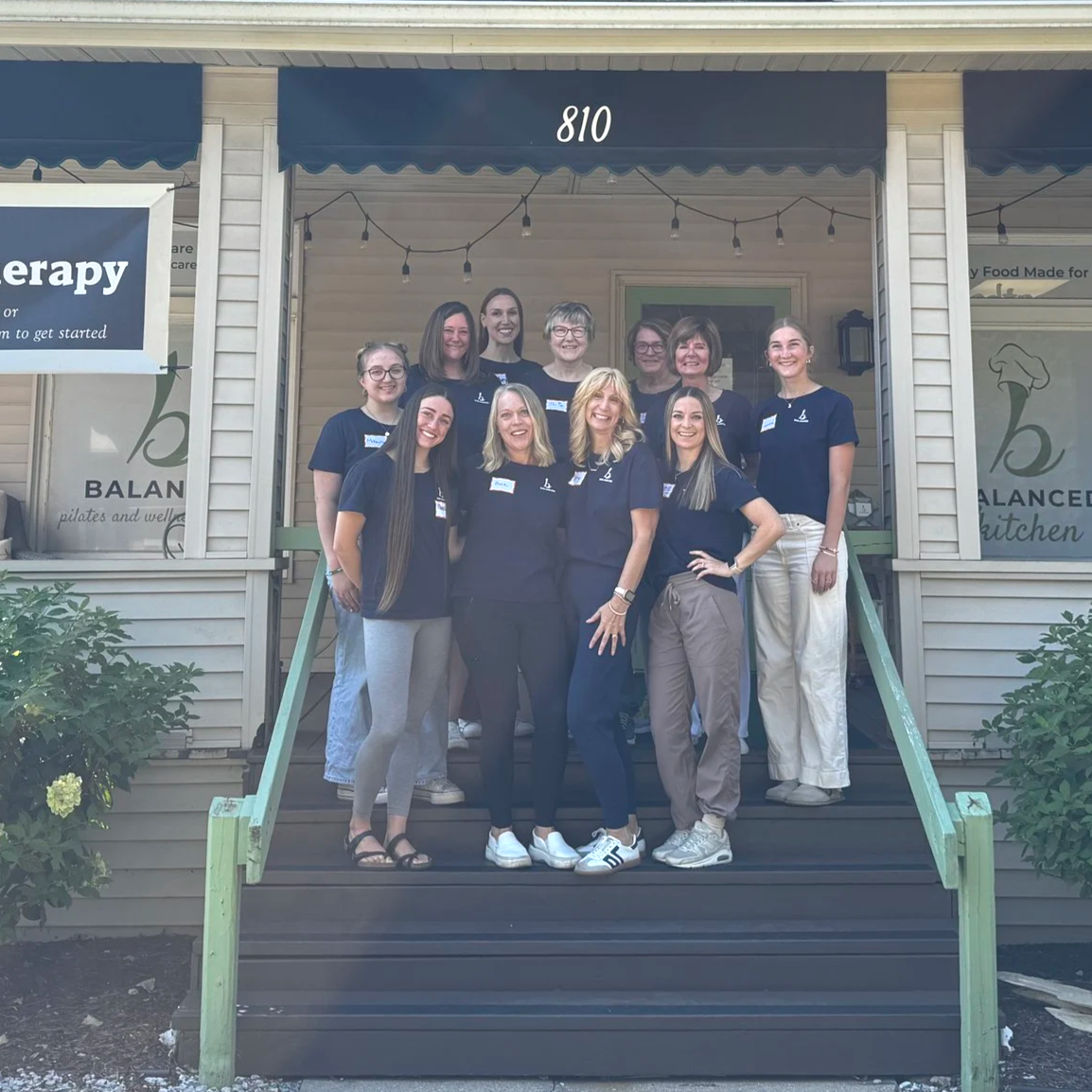 Group of women standing on the front porch of a building at 810 Ashman St in Midland, Michigan, smiling and posed for a photo, wearing matching navy blue shirts with a logo.