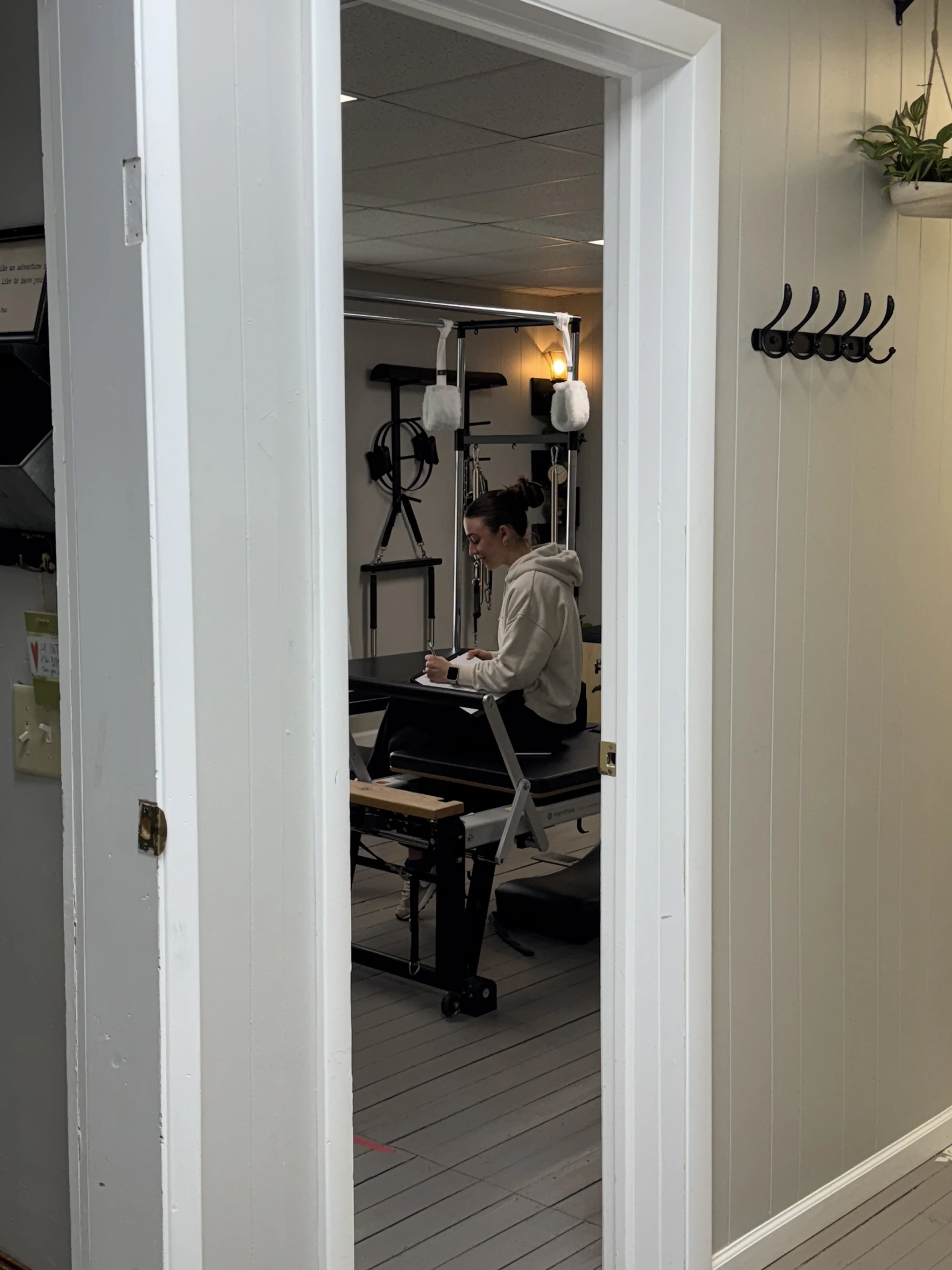 A woman sitting on a black exercise table in a small room, smiling and looking at her phone, with exercise equipment hanging on the wall behind her, illuminated by a warm wall light, and the room has wood plank flooring and gray walls.