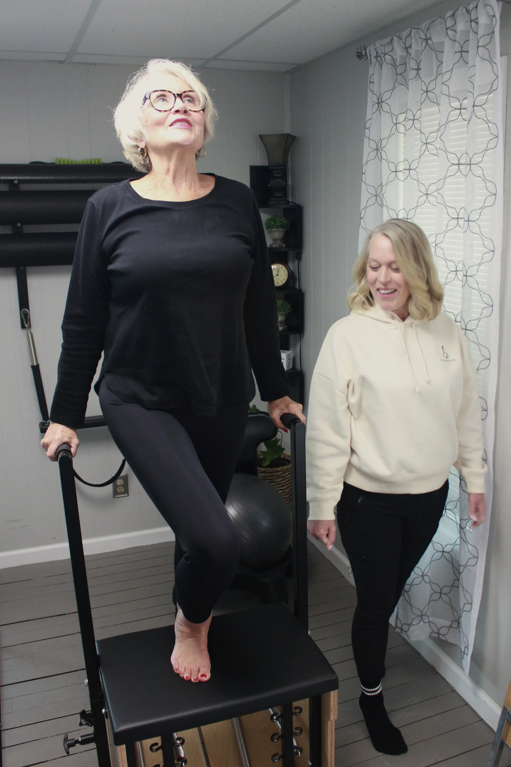 An older woman exercises on a treadmill with the assistance of a woman wearing a beige hoodie in a room with white walls and curtains.