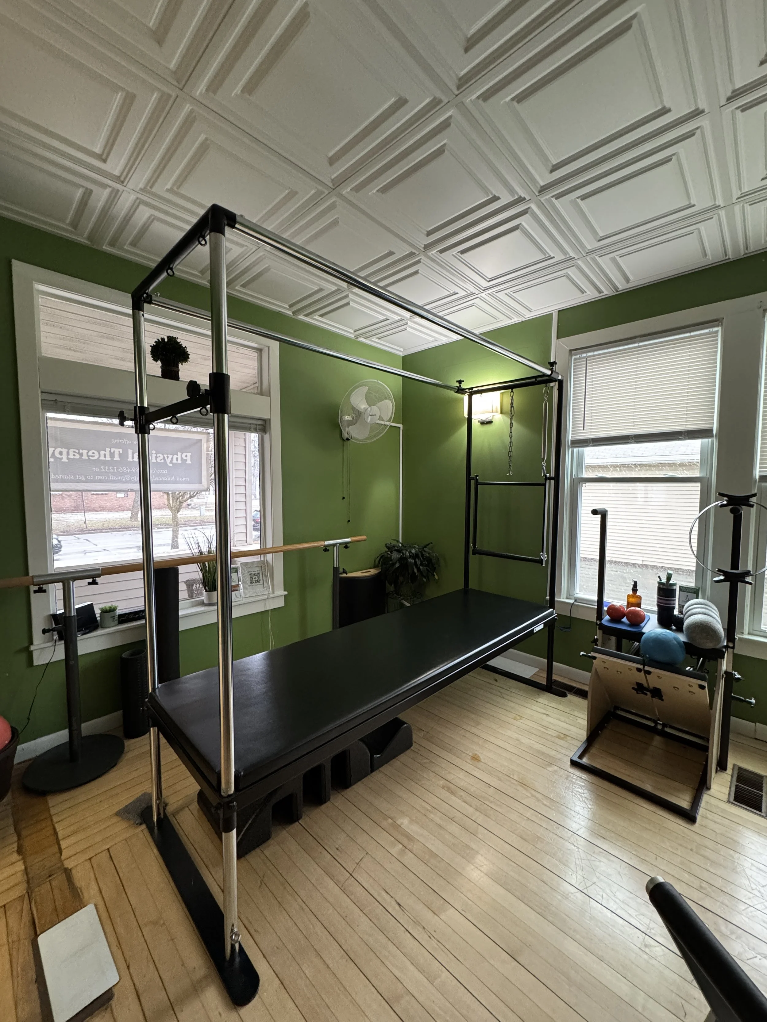 Private room with a black Pilates reformer, metal frame, and various exercise equipment near windows with blinds, green walls, and wood flooring at Balanced Pilates and Wellness Boutique in Midland, Michigan.