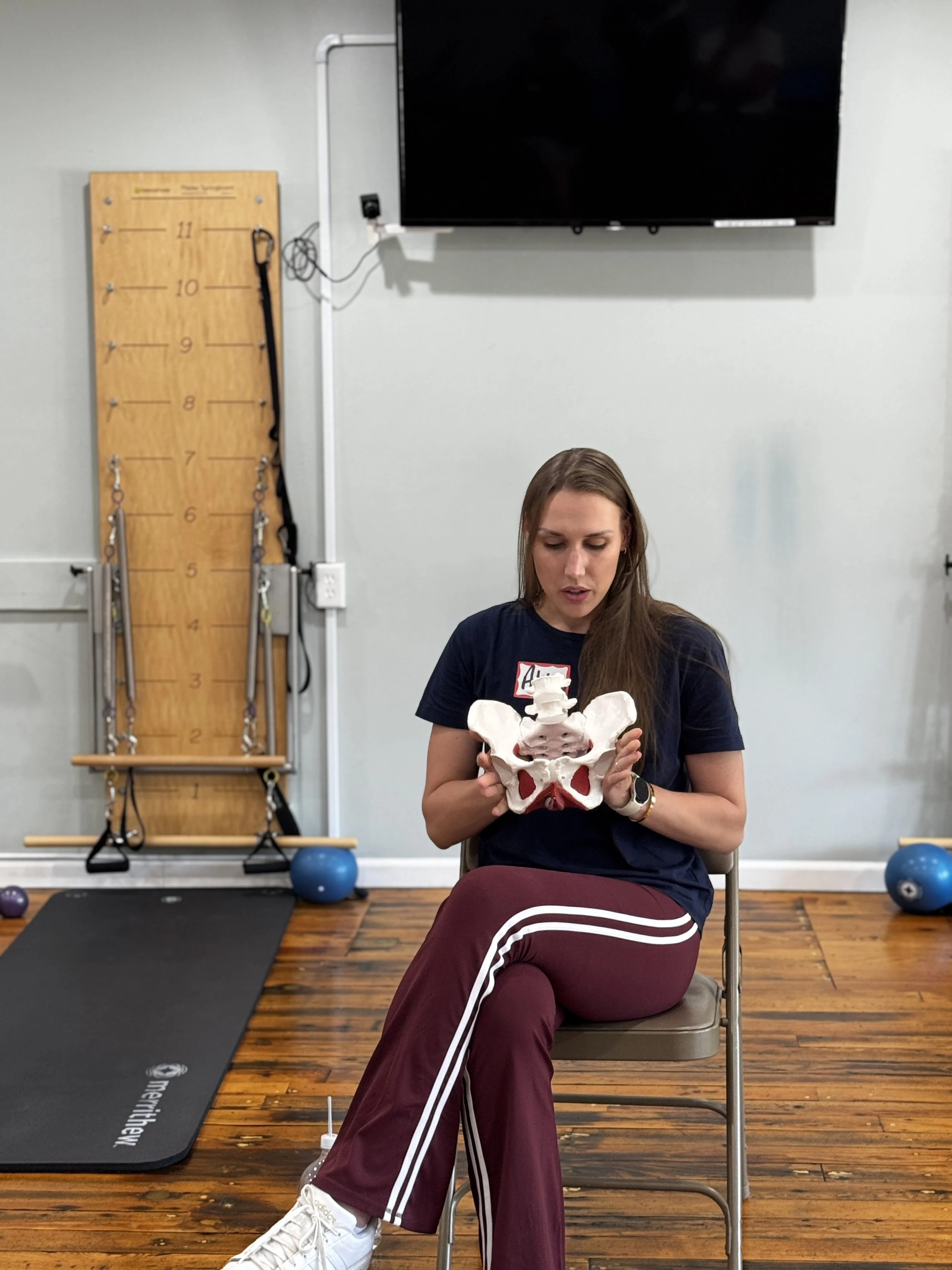 A woman sitting on a chair holding a model of a human pelvis and lower spine in a fitness or therapy room.