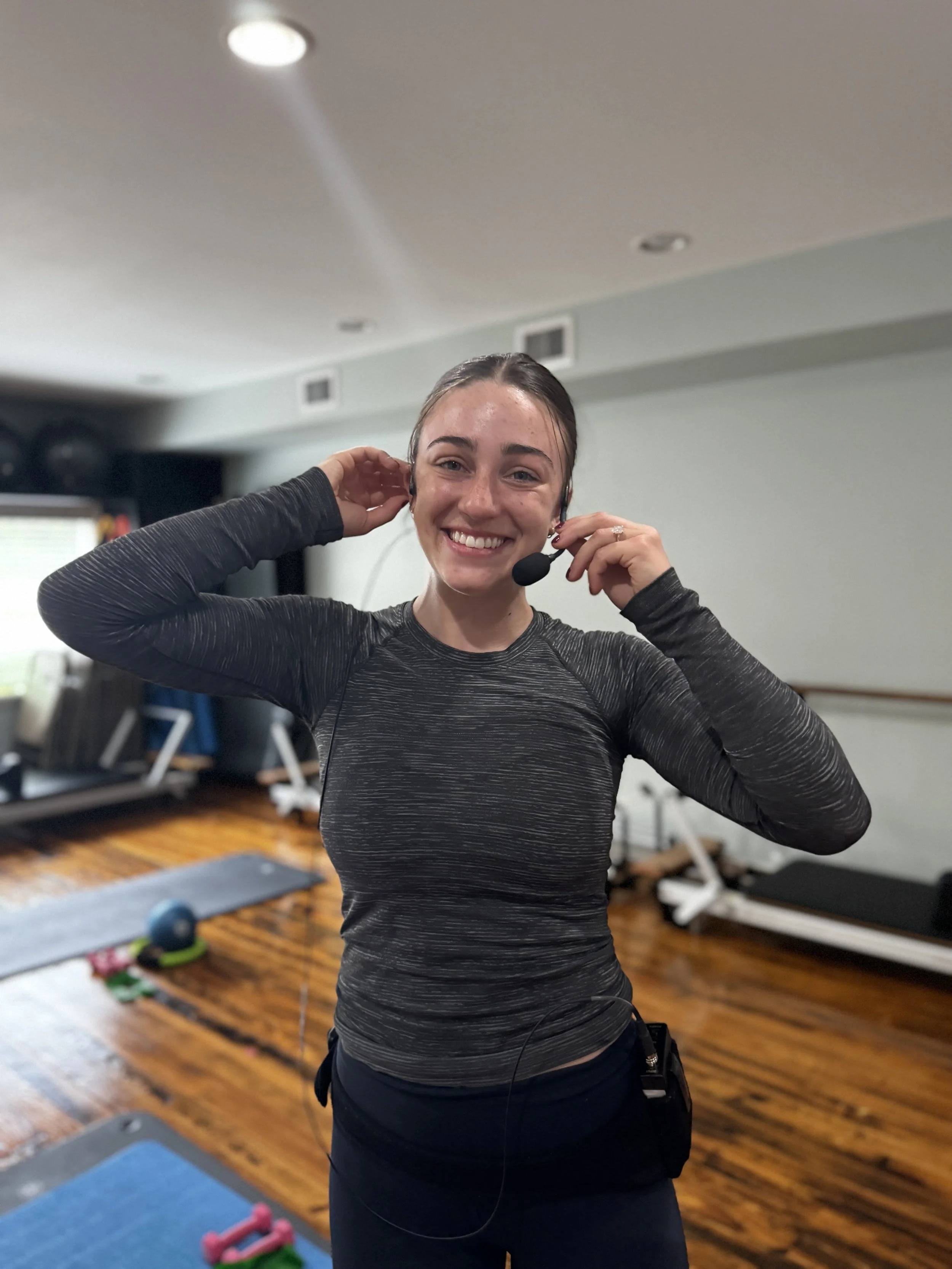 An instructor and adjusting her wireless headset in a Pilates studio in Midland, Michigan. She is wearing a gray, long-sleeved athletic top and black pants, with workout equipment and a wooden floor in the background.