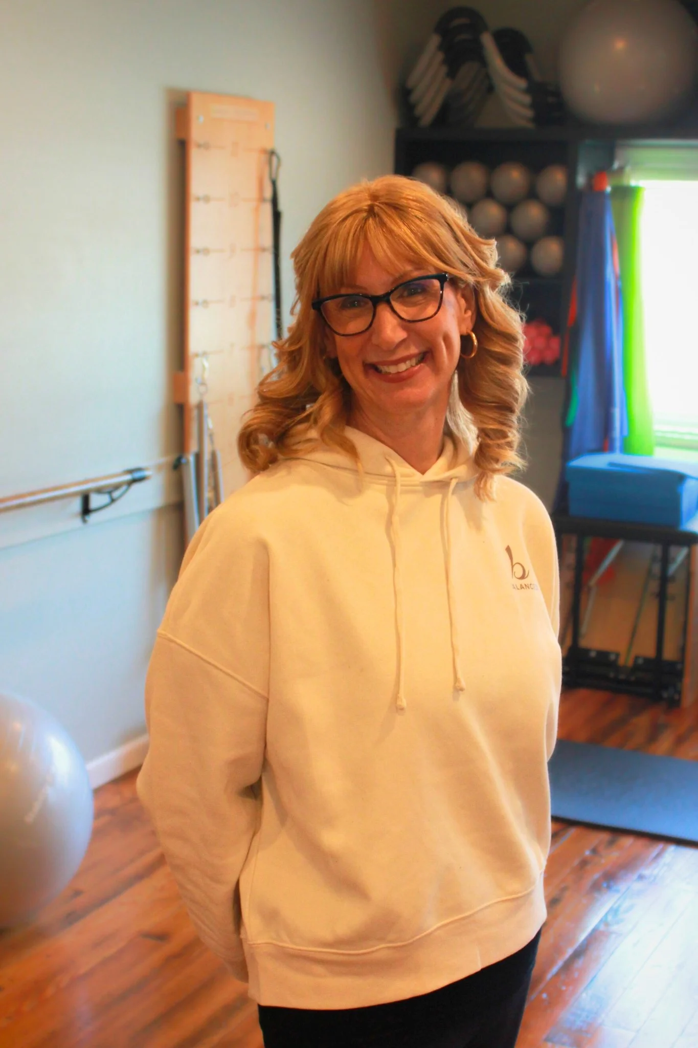 A woman with blonde hair, glasses, and hoop earrings smiling in a fitness room with exercise balls and equipment in the background.