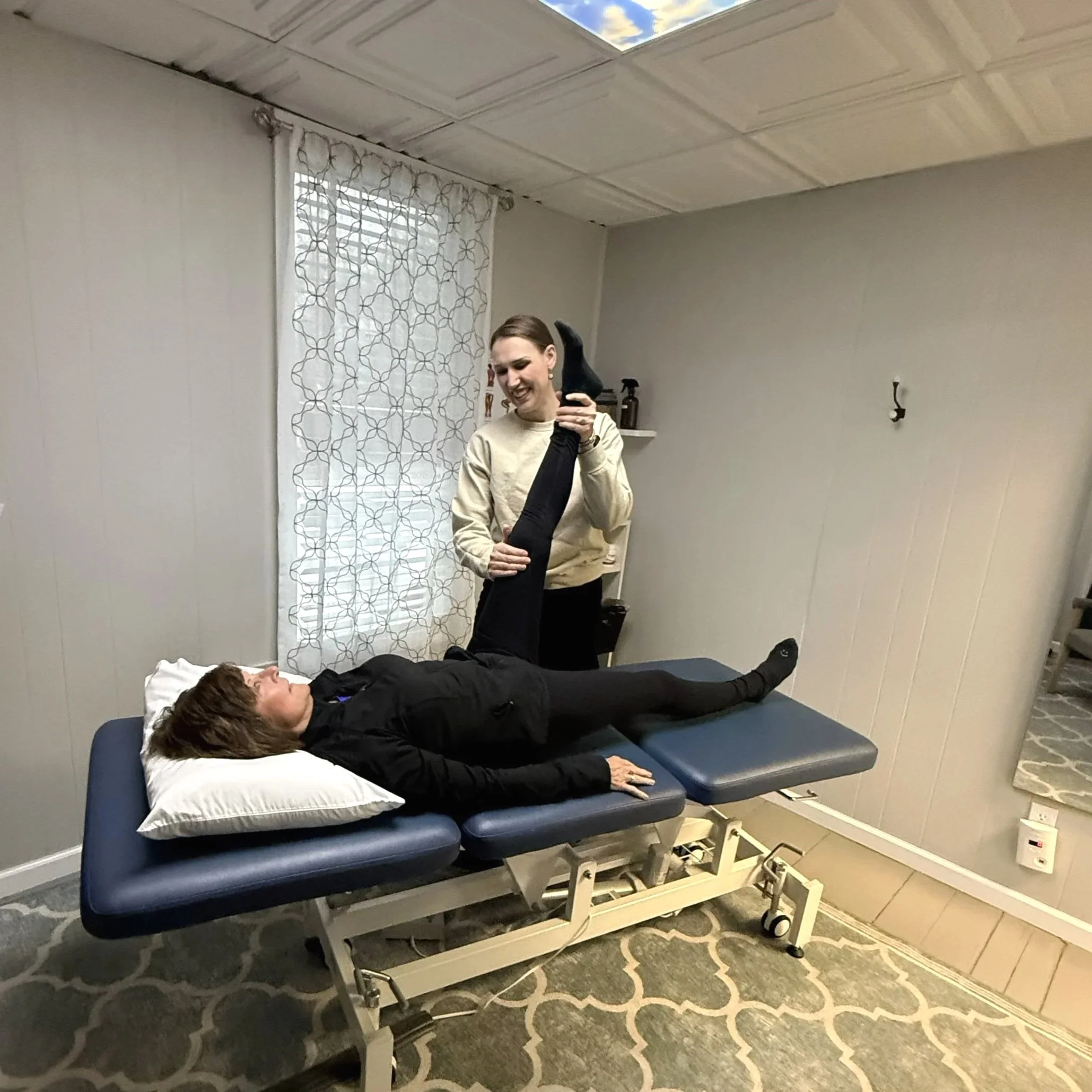 A woman is lying on a medical examination table with her eyes closed, receiving treatment from a physical therapist who is attending to her leg. The room has light-colored walls, a window with a patterned curtain, and a patterned rug on the floor.