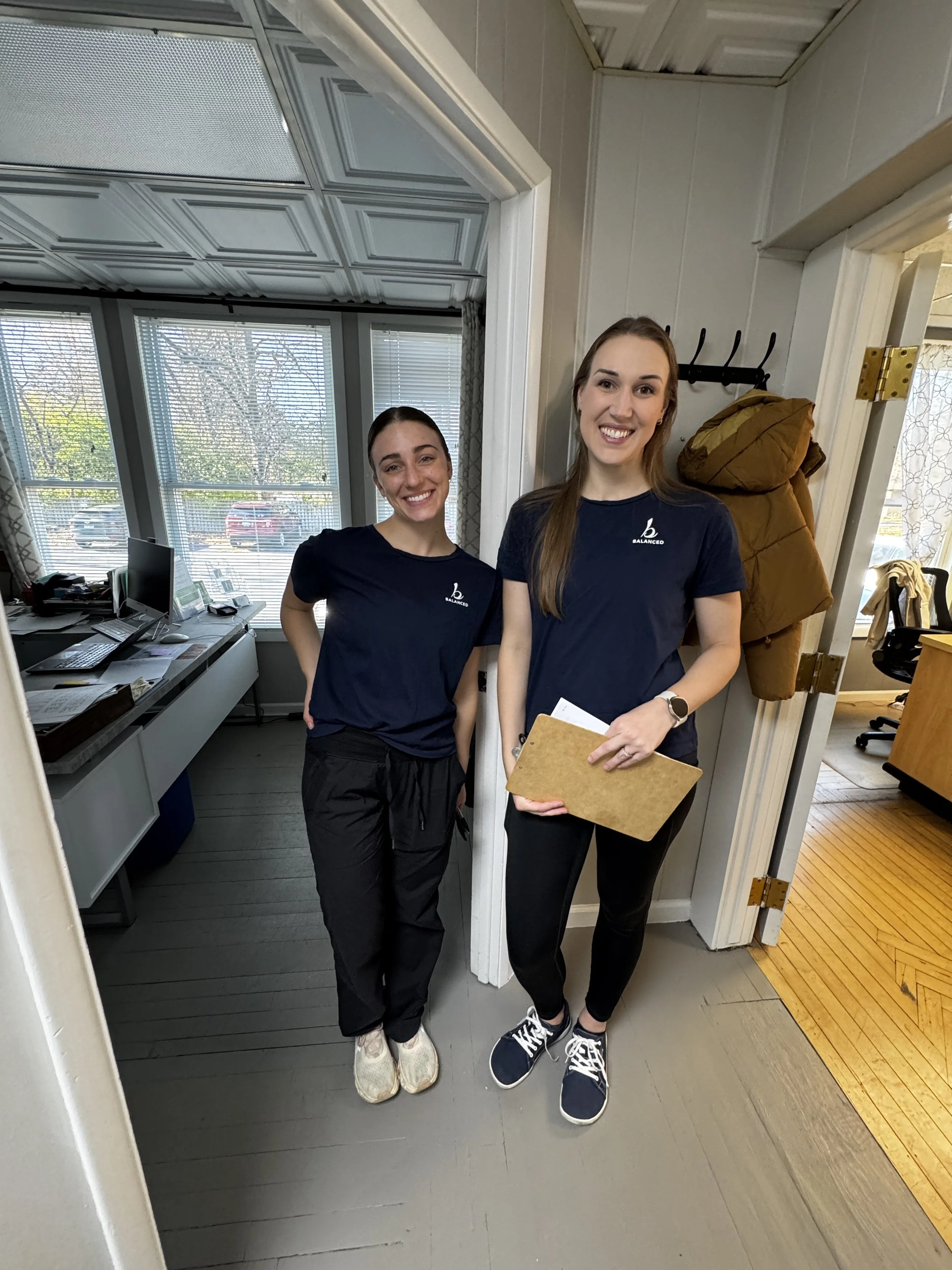 Two young women standing inside a room, smiling at the camera. The woman on the left has dark hair tied back, wearing a navy t-shirt and black pants. The woman on the right has long light brown hair, wearing a navy t-shirt with a logo, black pants, a