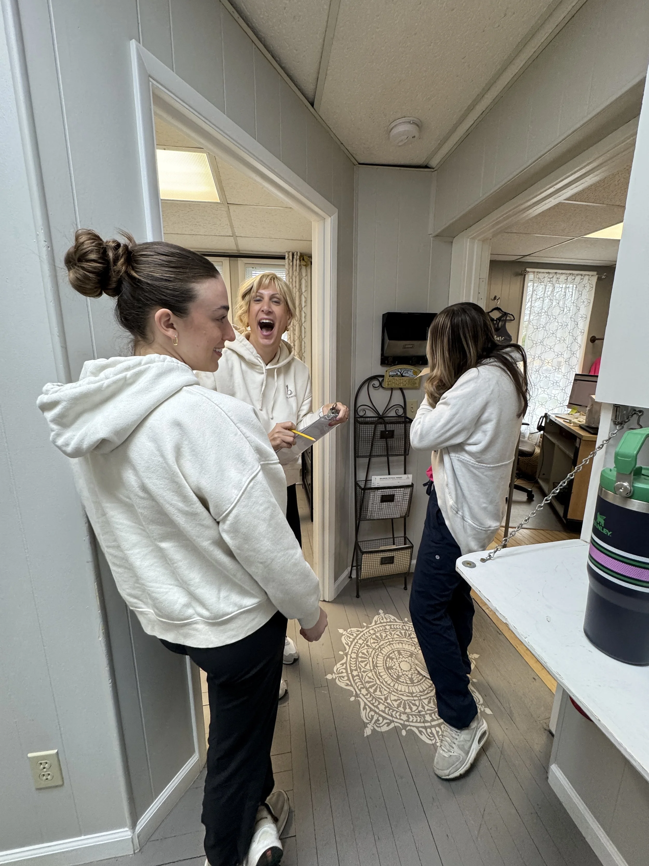 Three women standing in a room, engaging in a lively conversation. The woman in the center is smiling and holding a notepad and pen, wearing a white hoodie. The woman on the left is facing away, also in a white hoodie, while the woman on the right ha
