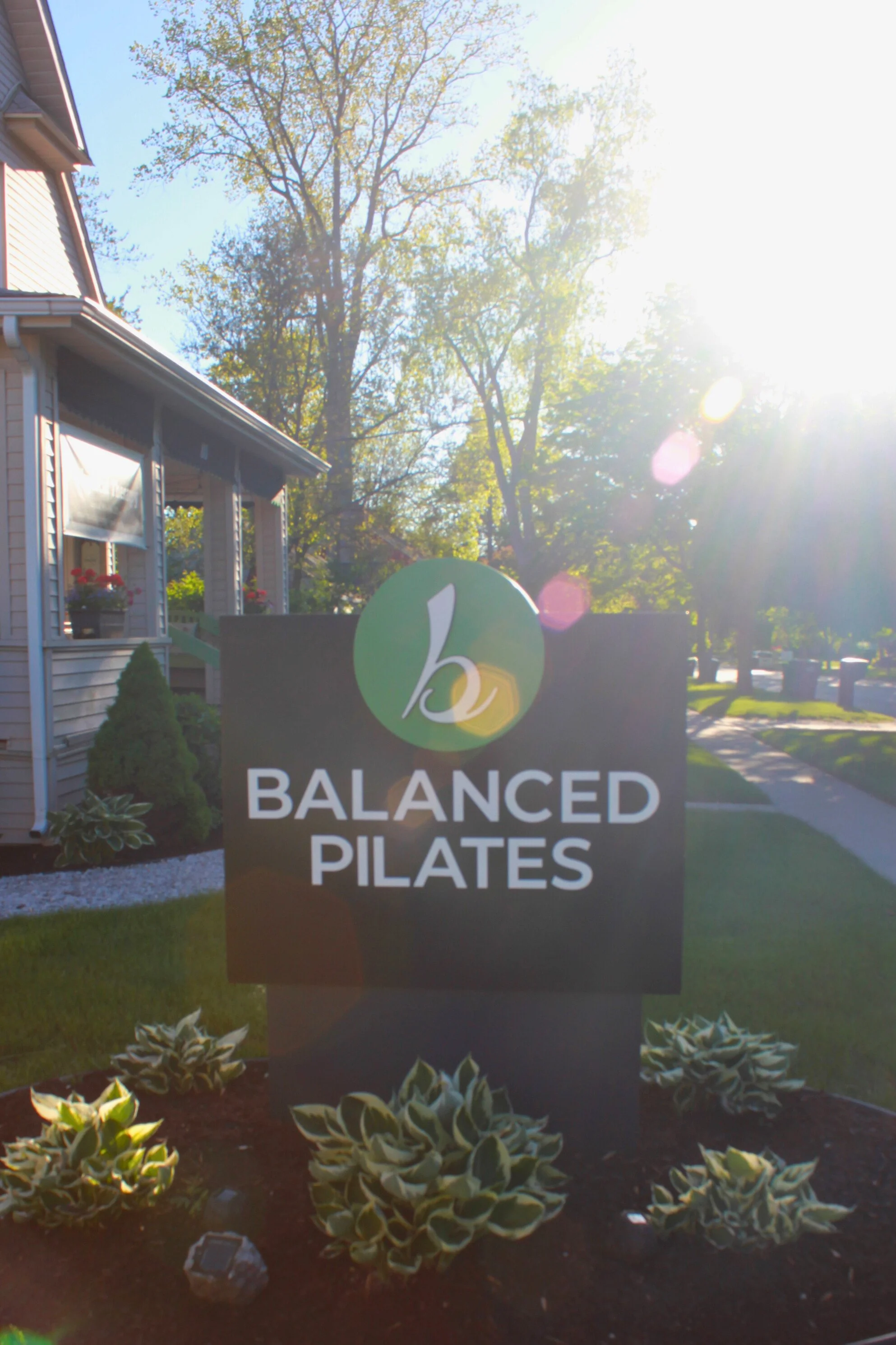 A sign for a business named 'Balanced Plates' in front of a house with trees in the background during daytime with sunlight.
