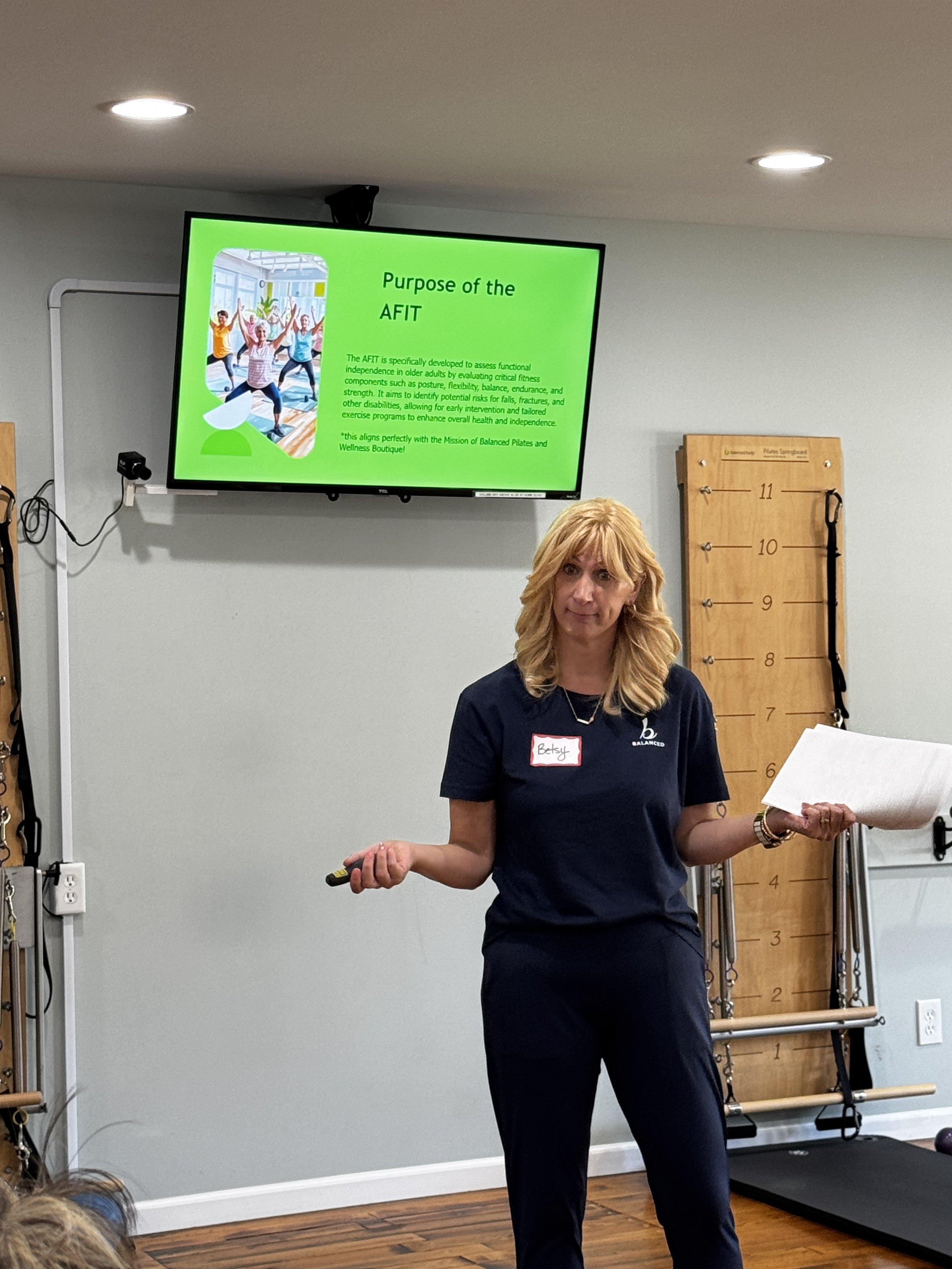 A woman with curly blonde hair wearing a dark blue t-shirt with a name tag and standing in front of a Pilates reformer machine, giving a presentation or demonstration with a remote in her right hand. There is a monitor on the wall behind her displayi