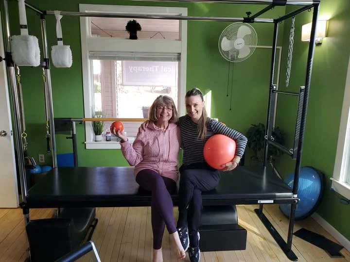 Two women sitting on a physiotherapy reformer exercise machine in a green room, smiling and holding therapy balls.