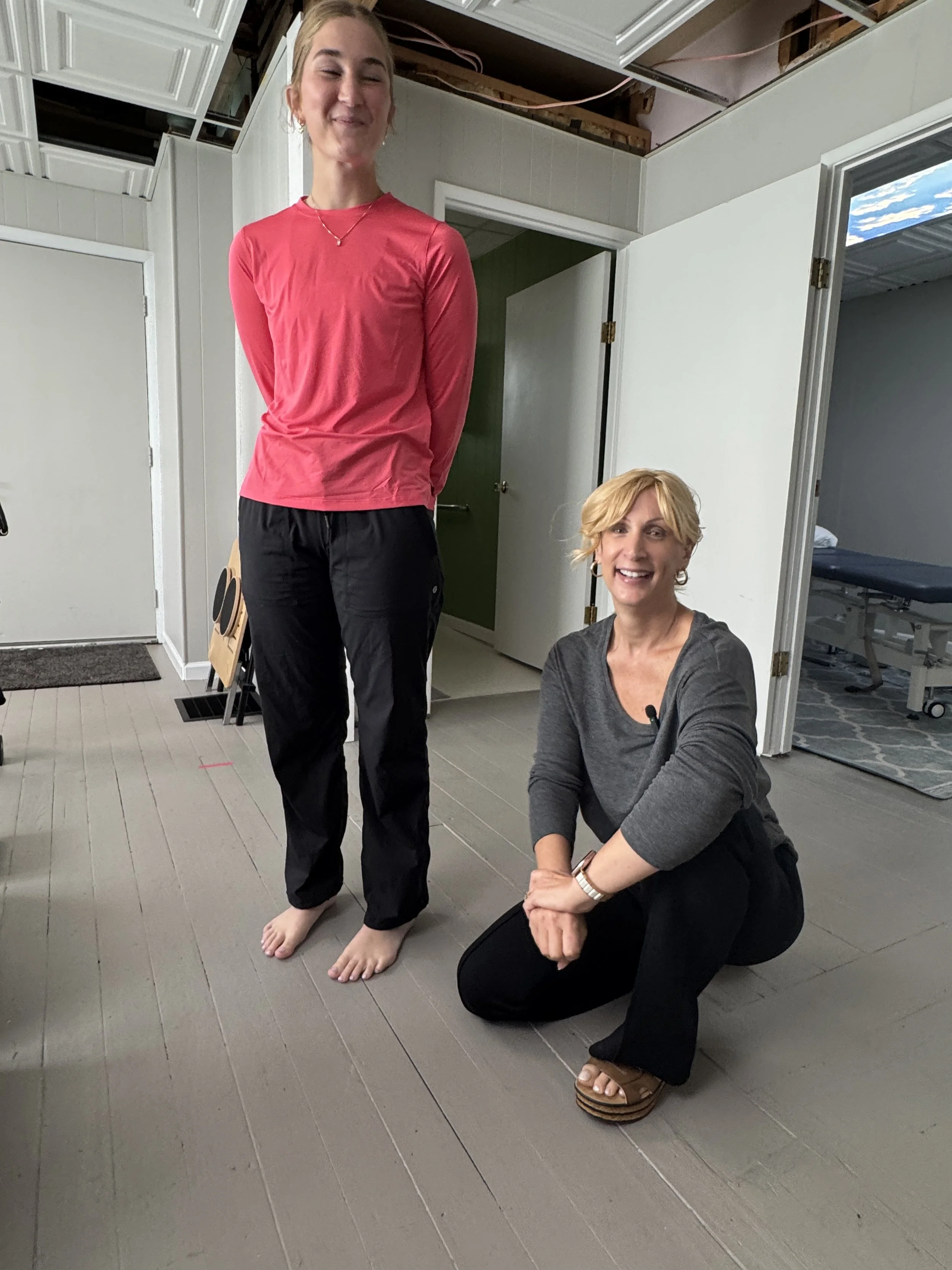 Two women in a room. One woman is standing with eyes closed, wearing a pink long-sleeve shirt and black pants. The other woman is kneeling on the floor, smiling, wearing a gray top and black pants, and is barefoot with sandals nearby. The room appear