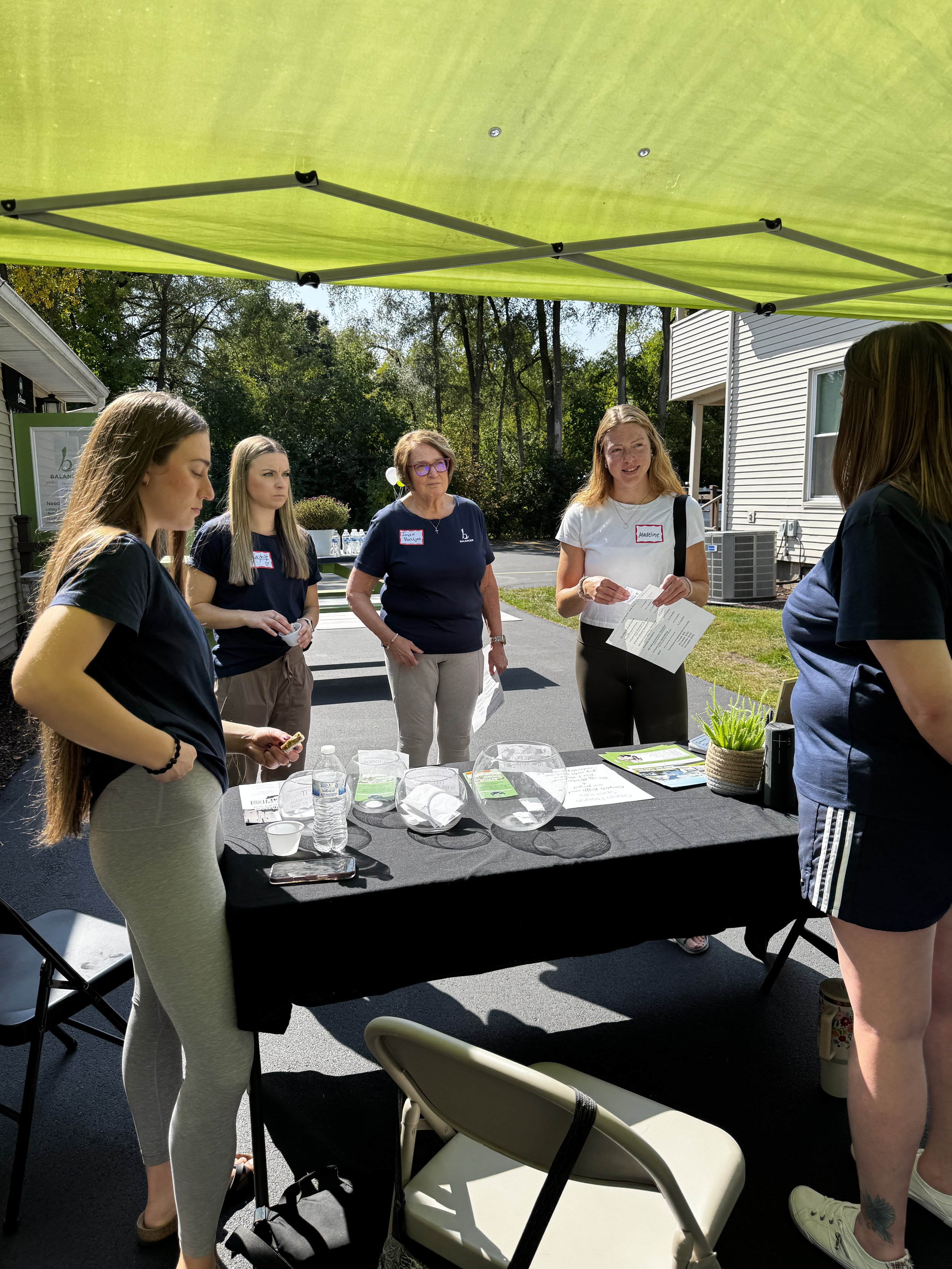 A group of women gathered outdoors around a table under a green canopy, engaging in conversation. The table is covered with informational materials and water bottles. There are trees and residential buildings in the background.