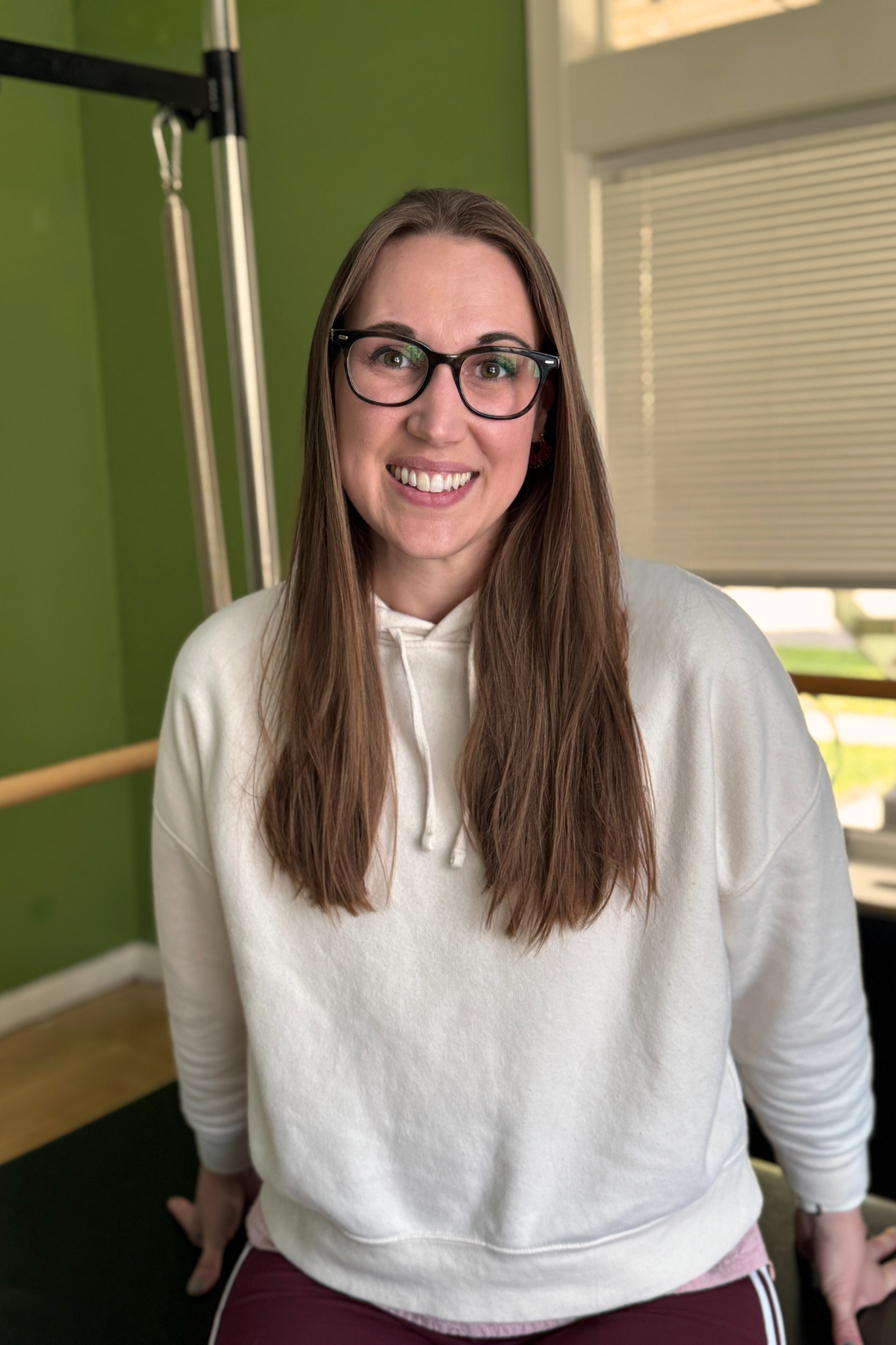 Dr. Abbey Thornock with long brown hair wearing glasses and a cream-colored sweatshirt in a private Pilates roomm.