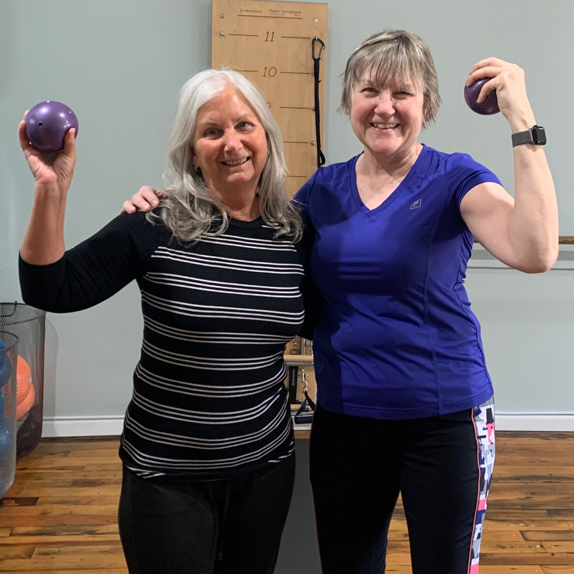Two women smiling and flexing their arms, holding purple exercise balls, at Balanced Pilates and Wellness Boutique in Midland, Michigan.