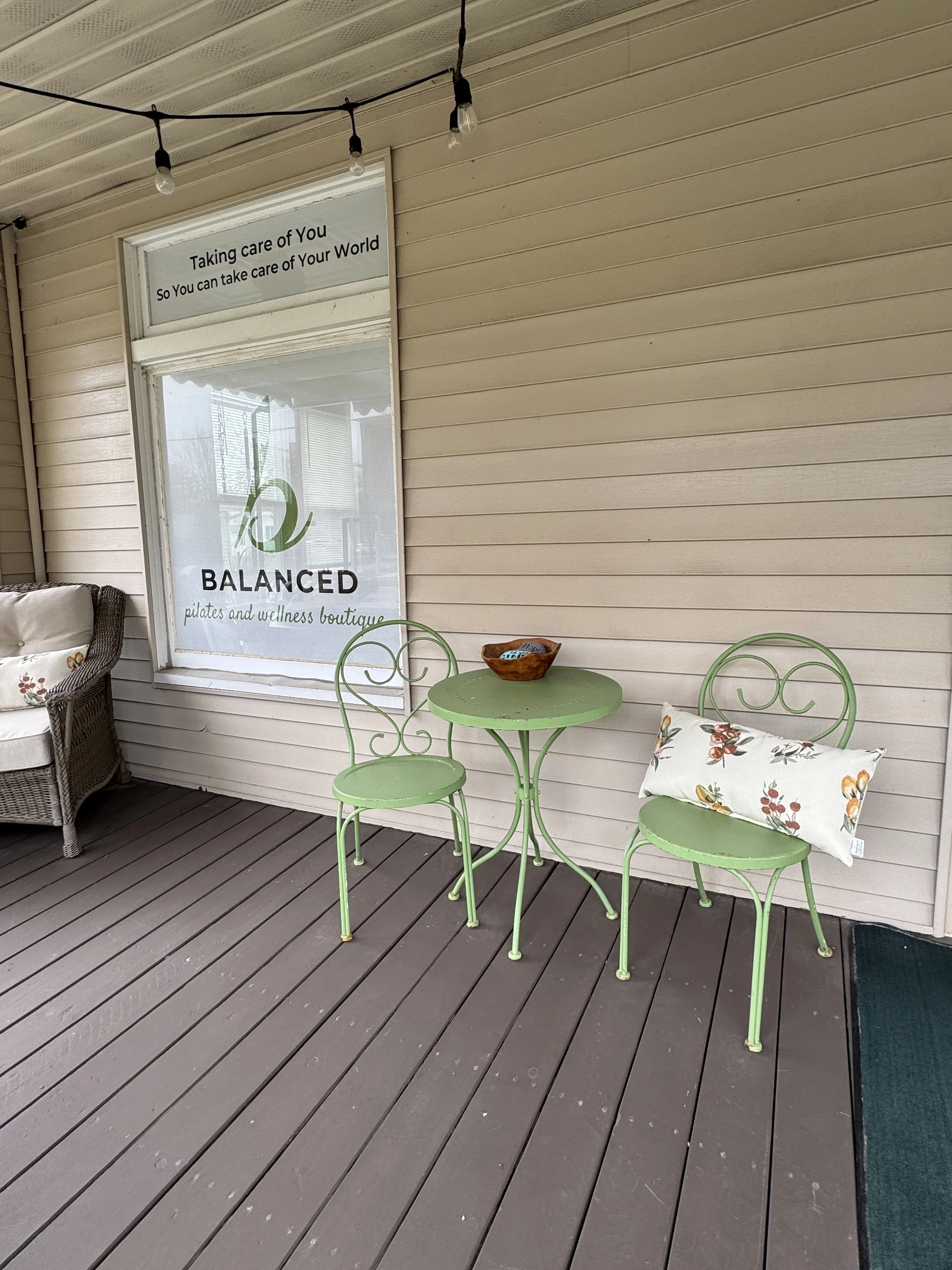A porch with a beige siding wall, a window with a sign saying "Taking care of You So You can take care of Your World," a wicker armchair with floral cushions, a small lime green round table, two matching lime green chairs, and a decorative pillow on 